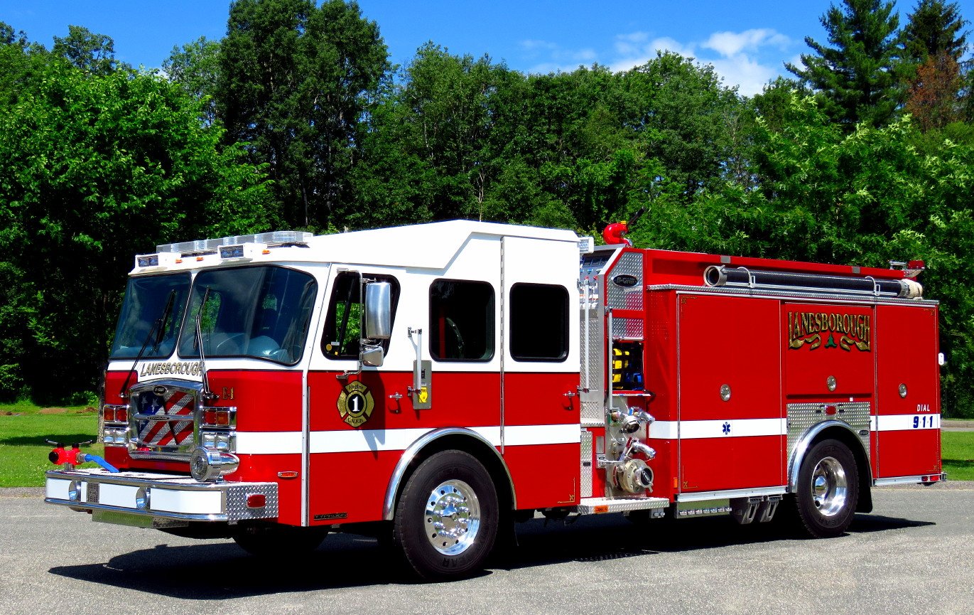 Red and white fire truck parked on pavement with green trees and blue sky in background.