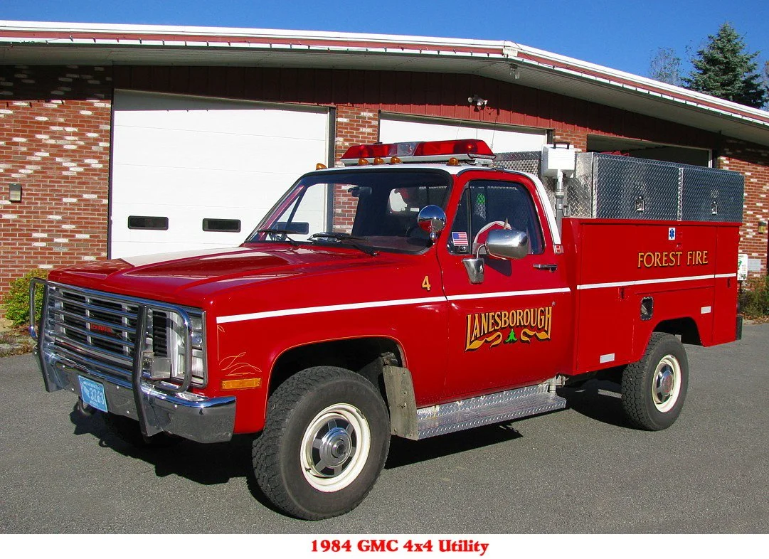 Red 1984 GMC 4x4 utility fire truck with 'Forest Fire' and 'Janesborough' written on the side, parked in front of a building with a white garage door.