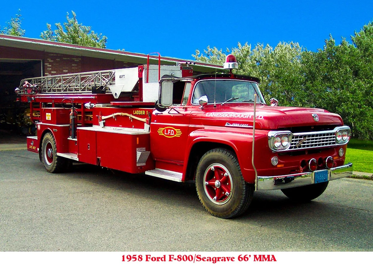 A vintage red 1958 Ford E-800 fire truck with a ladder on top, parked outdoors with greenery and a building in the background.