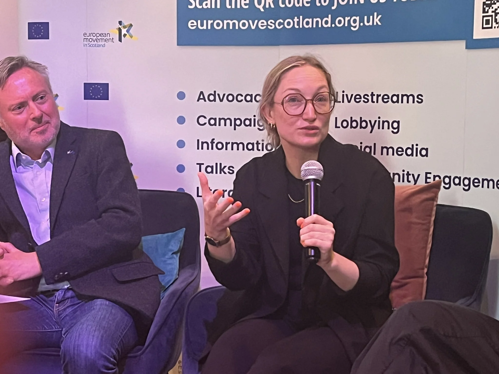 Woman speaking into a microphone at a panel discussion, with a man seated beside her, in front of a booth with text about advocacy, livestreams, campaigns, lobbying, and social media, and a banner for Euromovescotland.org.uk.