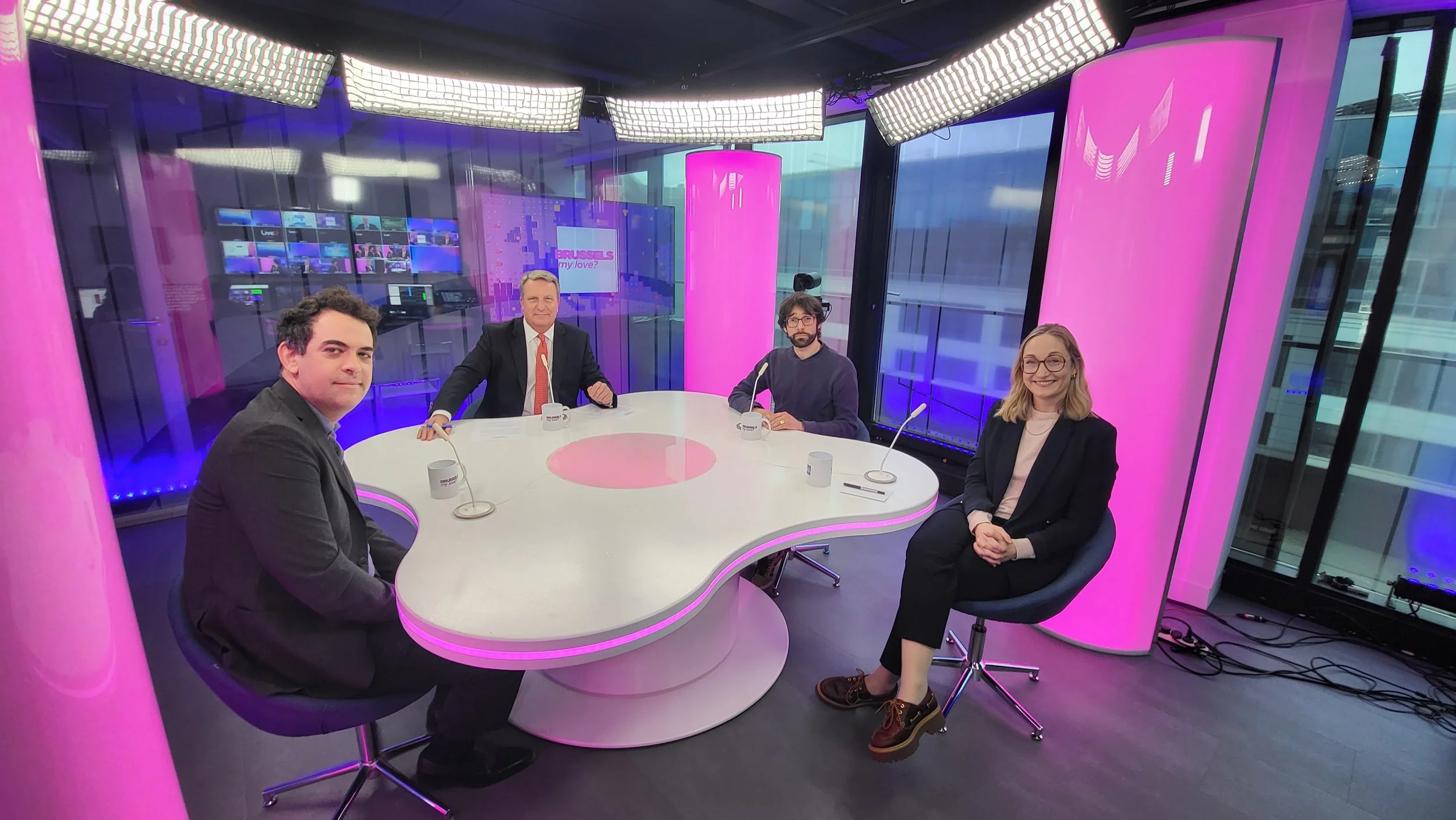 Four people sitting on chairs around a white, pink-lit table in a television studio with pink columns and large windows. The studio features multiple monitors and lighting equipment in the background.