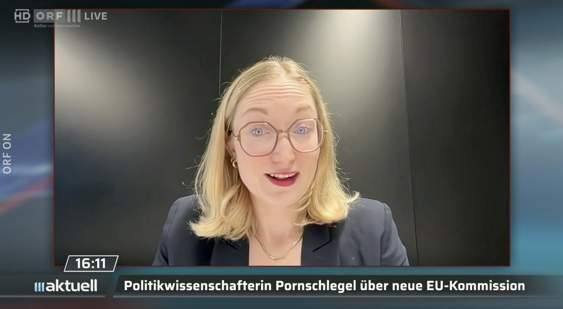 A woman with blonde hair and glasses appearing on a news broadcast, with the headline in German about a political scientist discussing the new EU Commission.