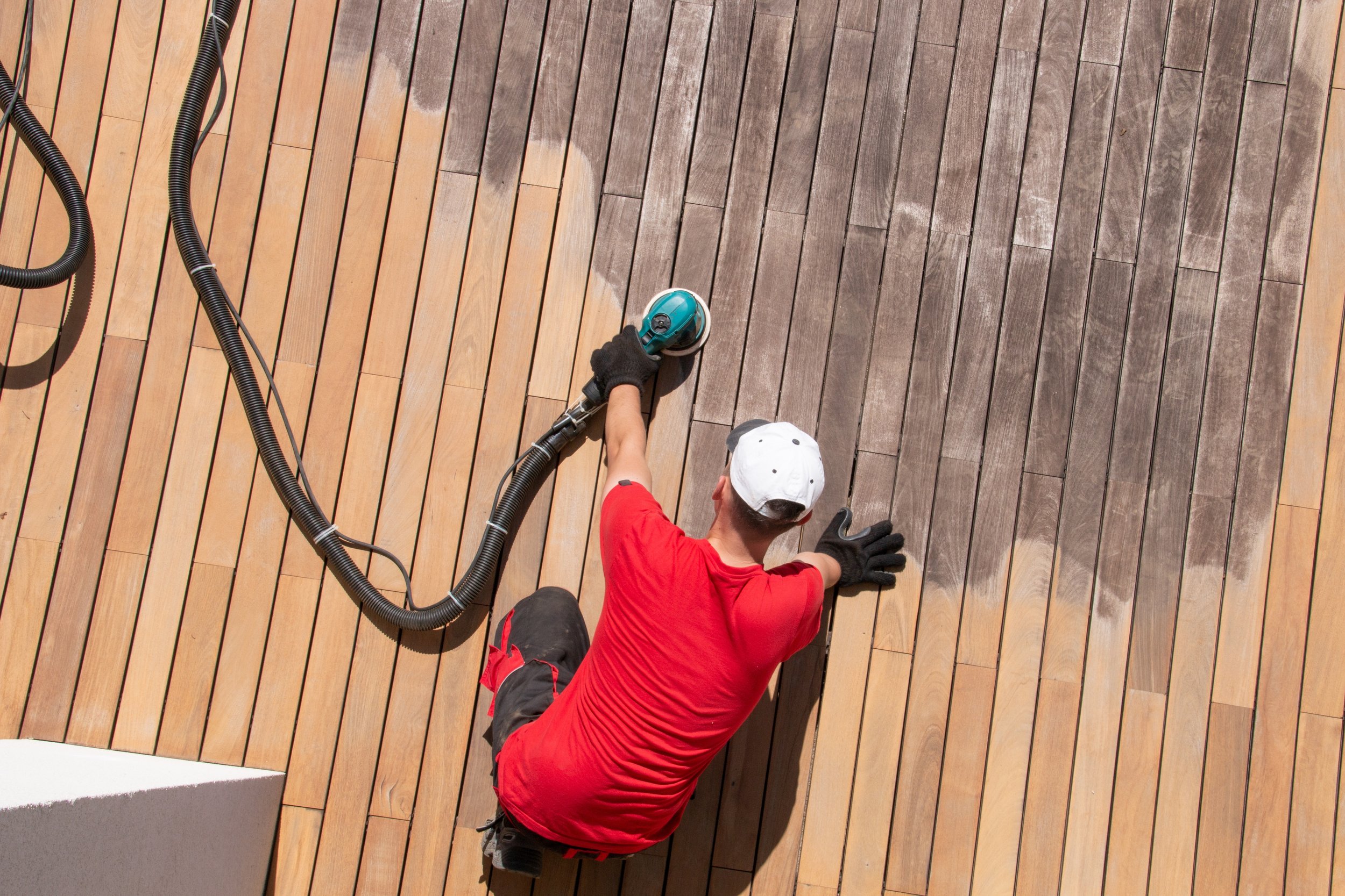 A person wearing a white cap, red shirt, black shorts, and black gloves is cleaning a wooden deck with a power washer.
