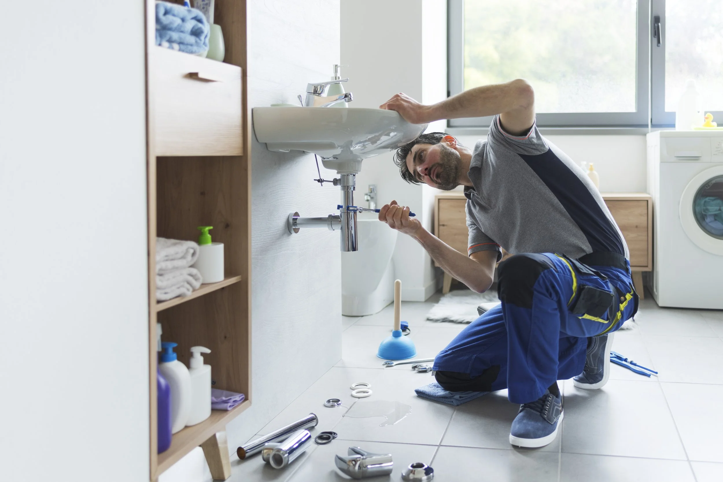 A man kneeling on the bathroom floor fixing a leaking bathroom sink with various tools and parts scattered around, in a bright bathroom with a washing machine and window.