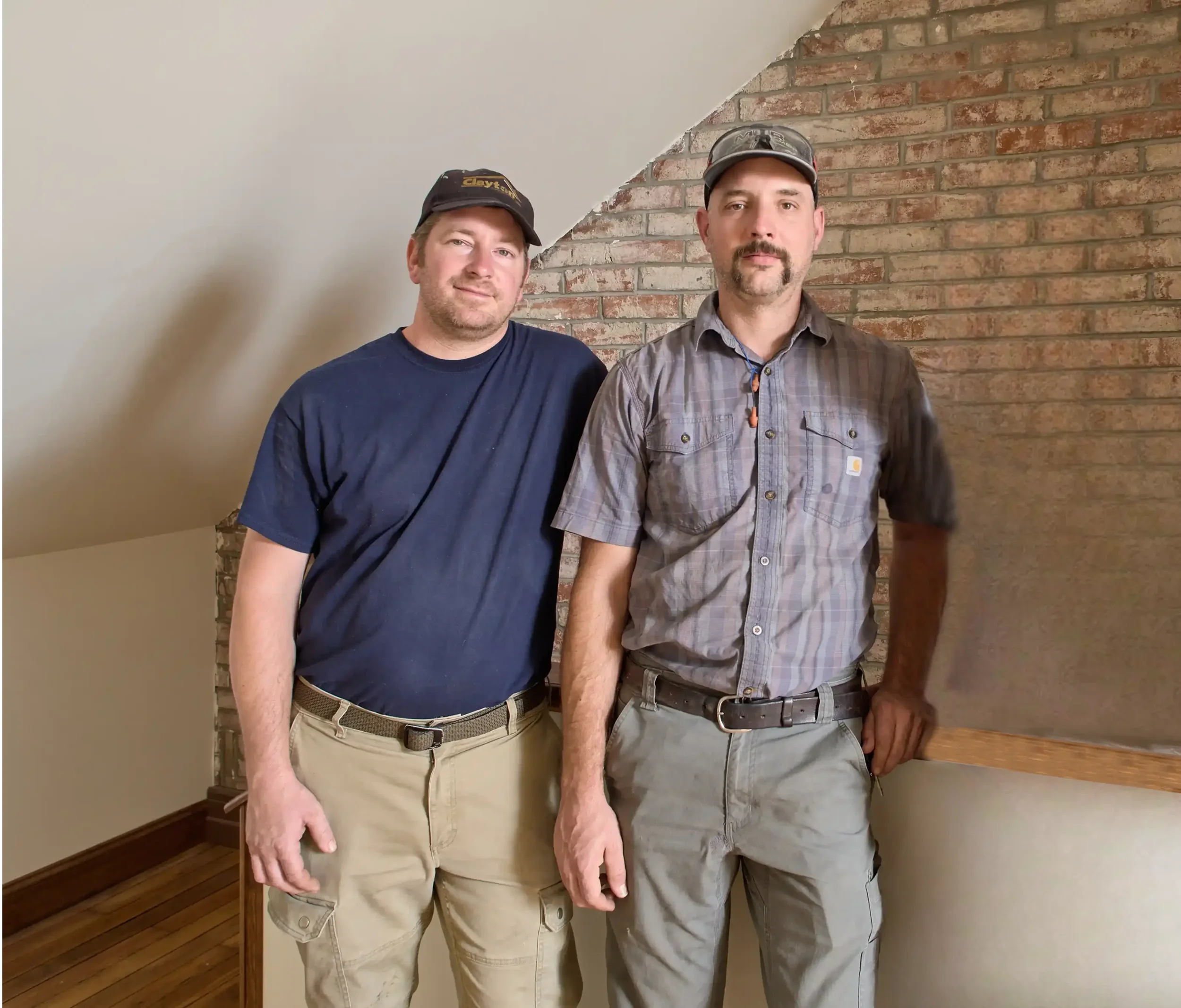 Two men standing indoors with a brick wall behind them. One man is in a navy t-shirt and khaki pants, and the other is in a gray plaid shirt and khaki pants, both wearing caps.