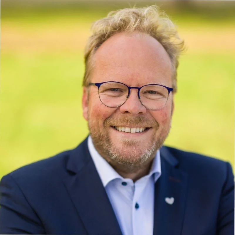 Black and white portrait of a man with short hair and a beard, wearing a blazer, standing in front of a brick wall.