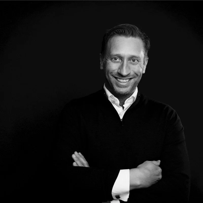 A bald man with a light complexion in a pink striped shirt outdoors with trees in the background.