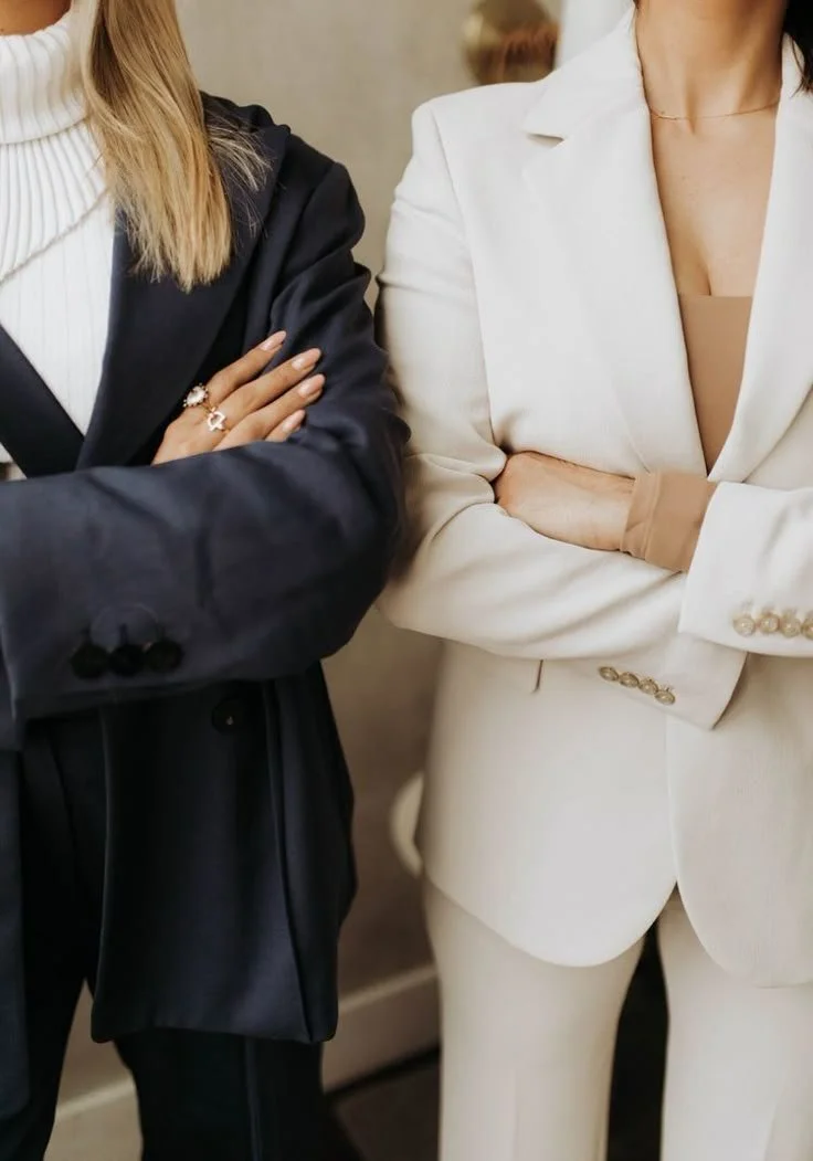 Close-up of two women standing side by side with their arms crossed, dressed in business attire, one in a black blazer and the other in a cream-colored blazer.