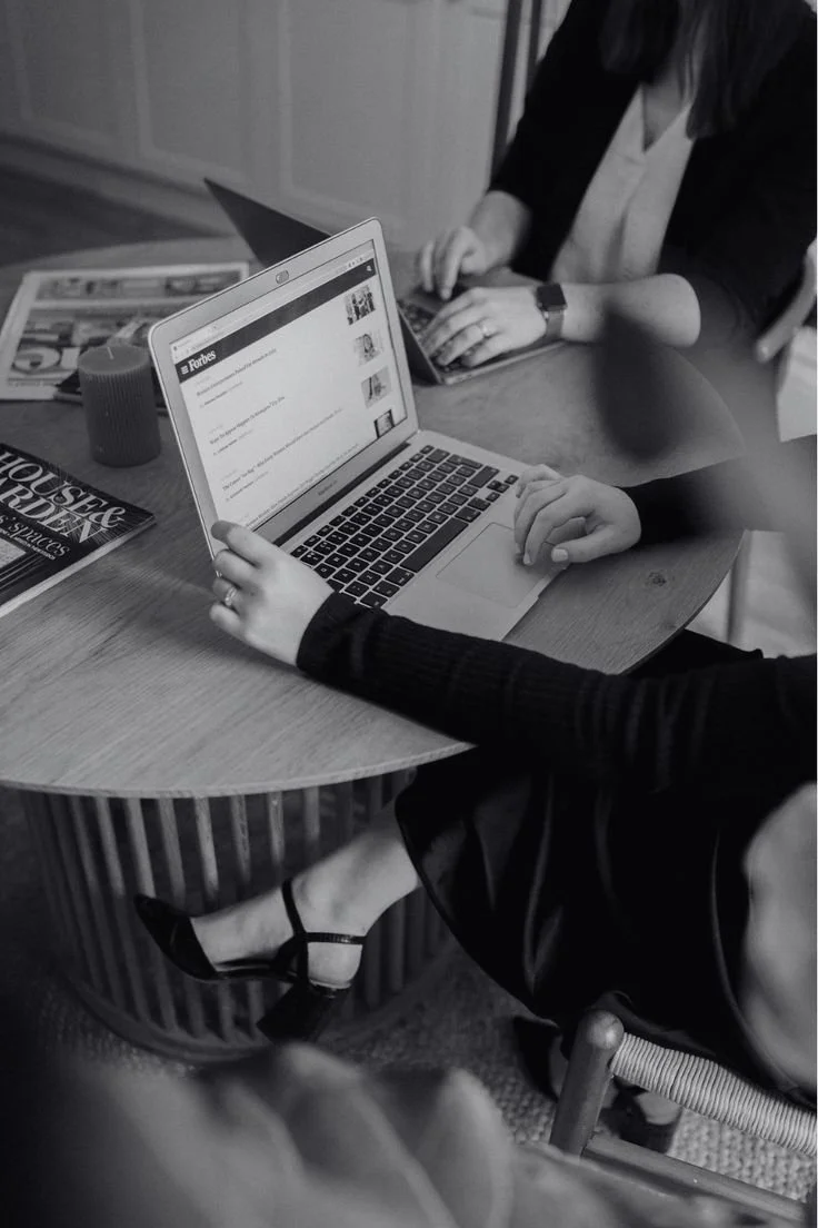 Two women working at a desk with various art supplies, a candle, and papers in front of a large window.
