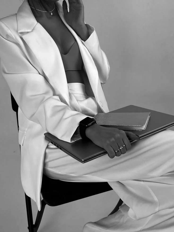 Two women smiling and laughing together, one in a white outfit and the other in a dark blazer, standing against a plain background.