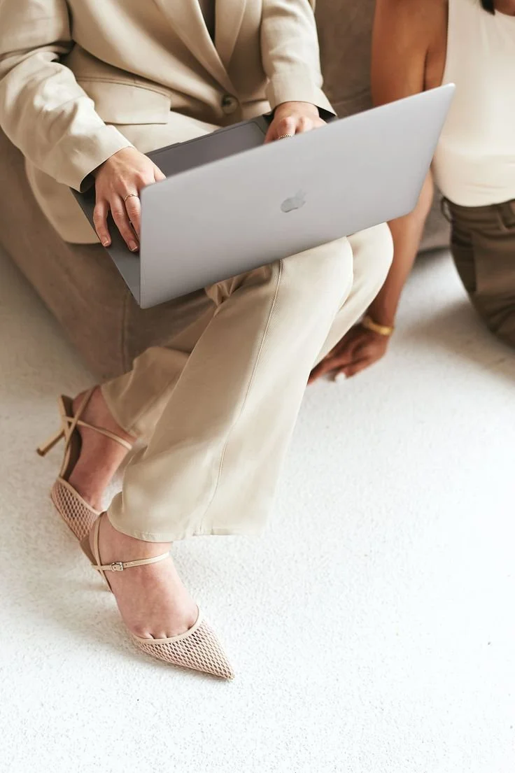 A woman in beige pants and high heels sitting on a couch, holding a silver MacBook laptop on her lap.