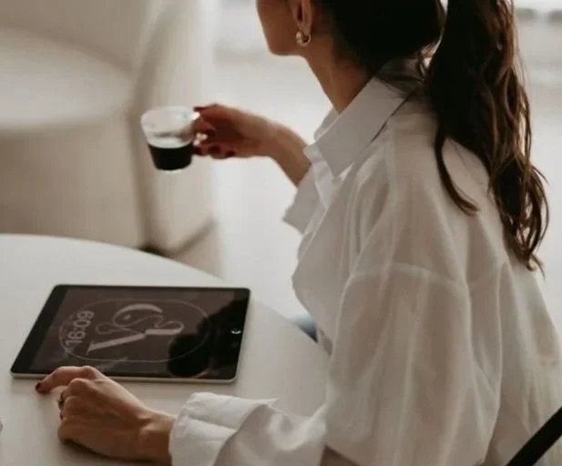 A woman sitting at a white table with a tablet and a small vape pen, holding a glass of dark liquid, in a bright room with large windows in the background.