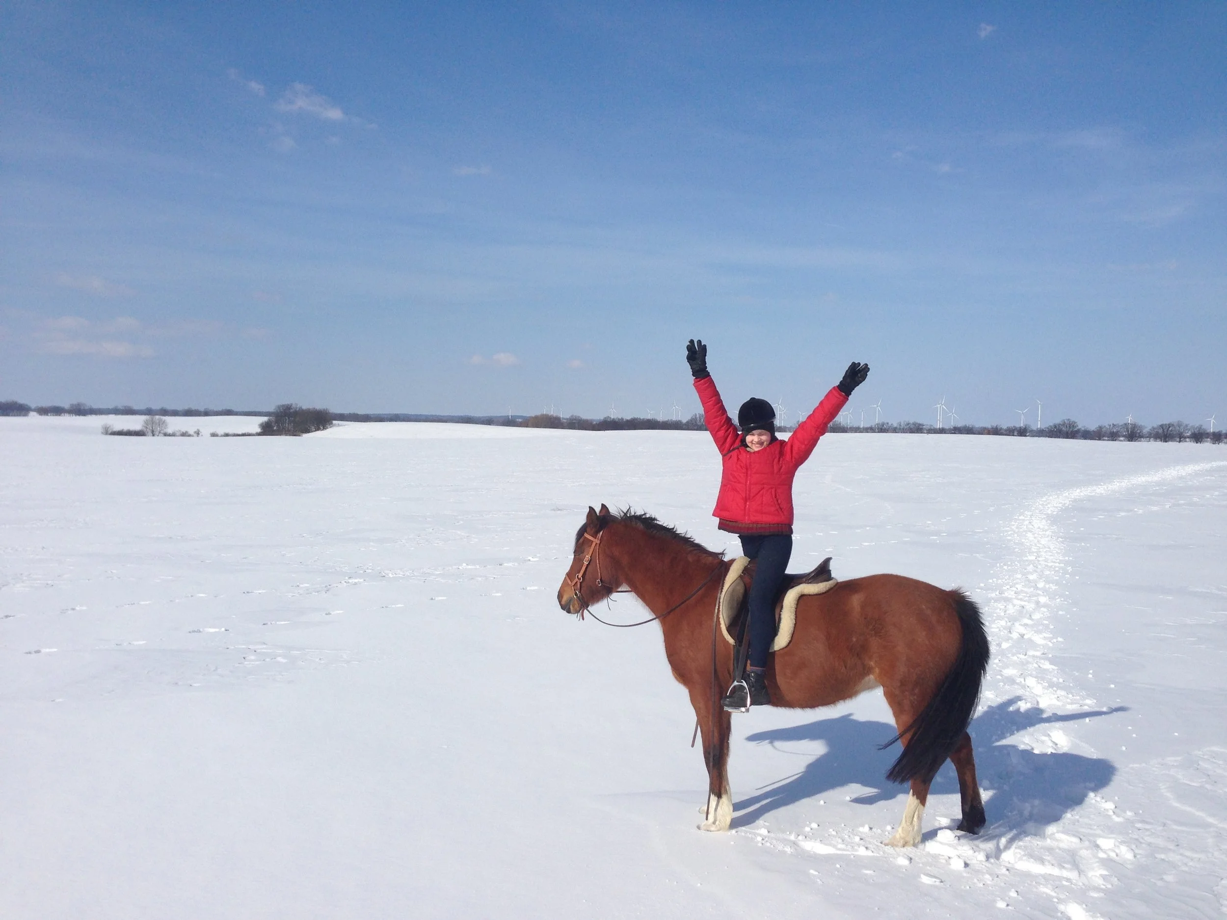 Frau reitet auf einem braunen Pferd im verschneiten Landschaft unter einem blauen Himmel mit Wolken und Windkraftanlagen im Hintergrund.