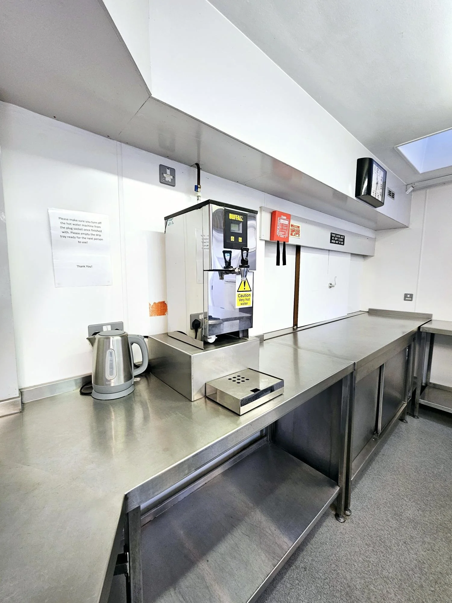 A stainless steel countertop with a juice dispenser, electric kettle, and a drip tray in a kitchen or break room.