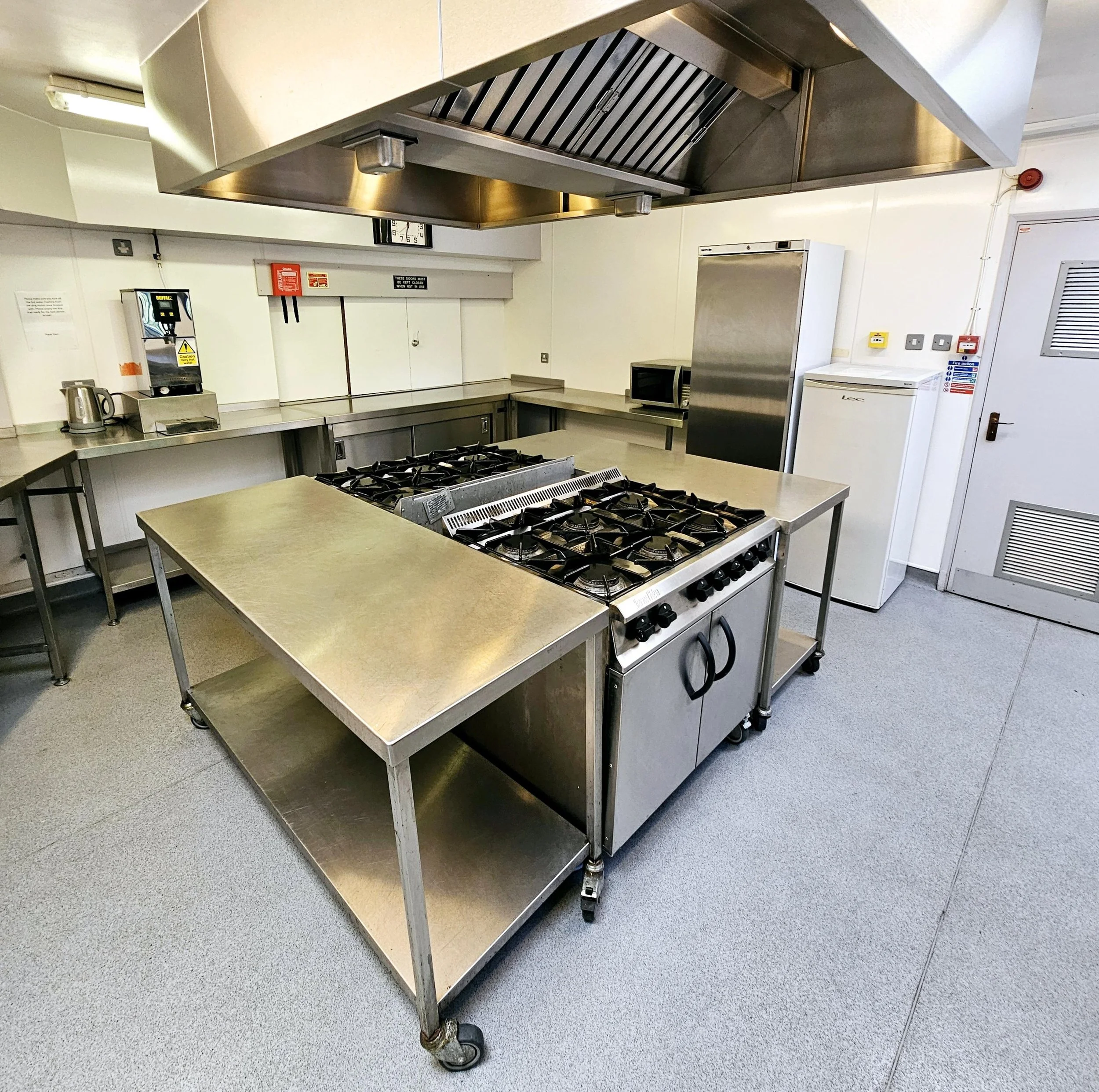 Empty commercial kitchen with stainless steel appliances, a stove, microwave, refrigerator, and various countertop surfaces.