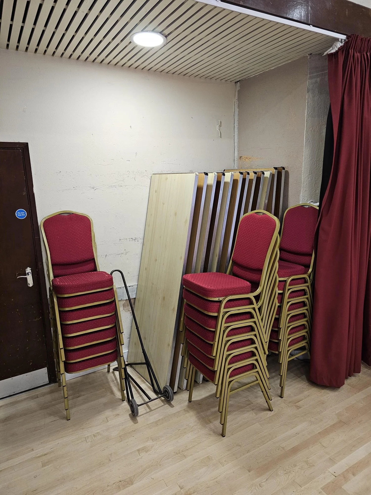 Stacked red chairs with gold frames, leaning against a wall, next to a black shopping trolley, with wooden panels and a red curtain nearby in an indoor space.