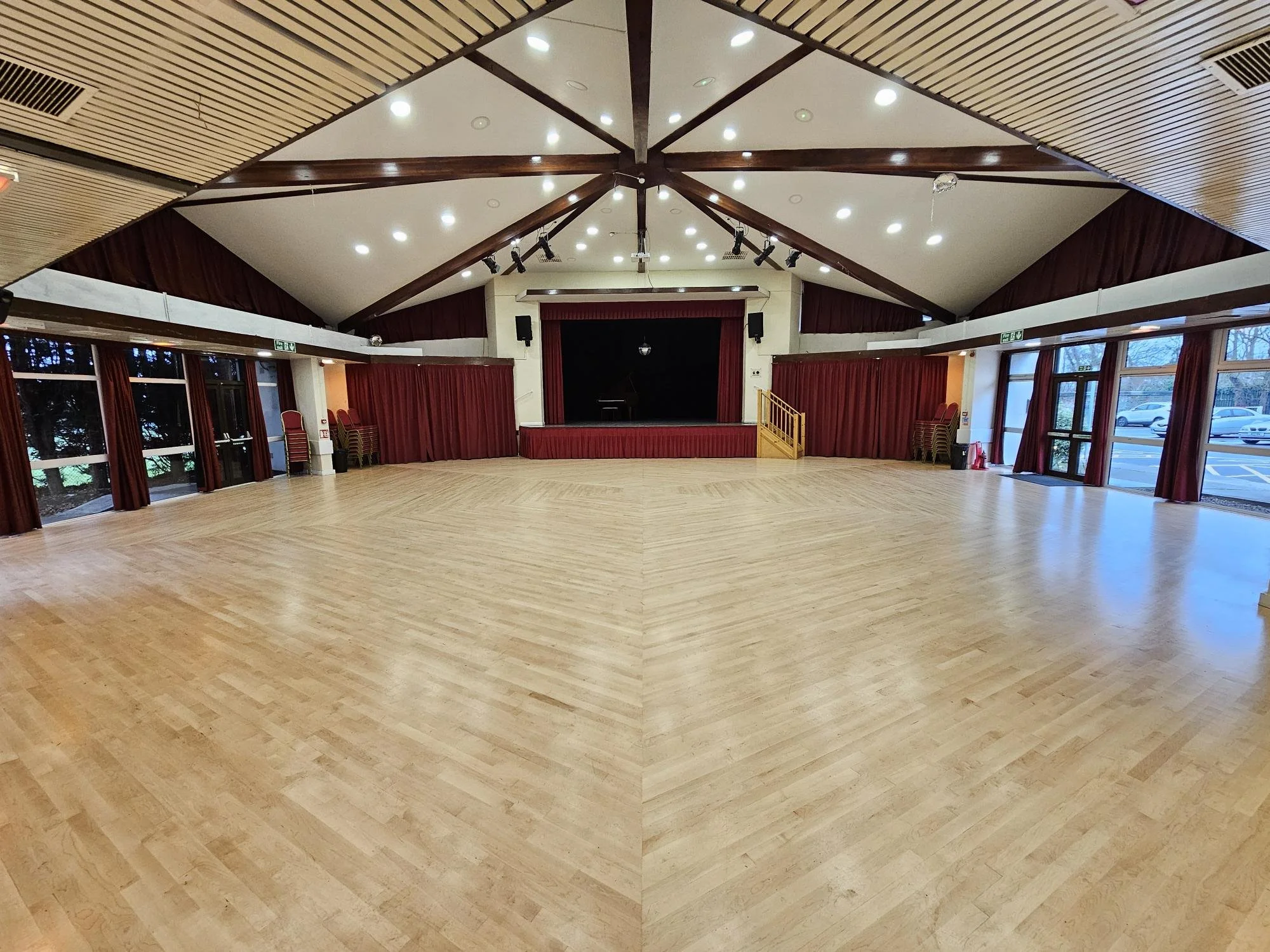 Large empty event hall with a stage, wooden flooring, red curtains, and ceiling lights.