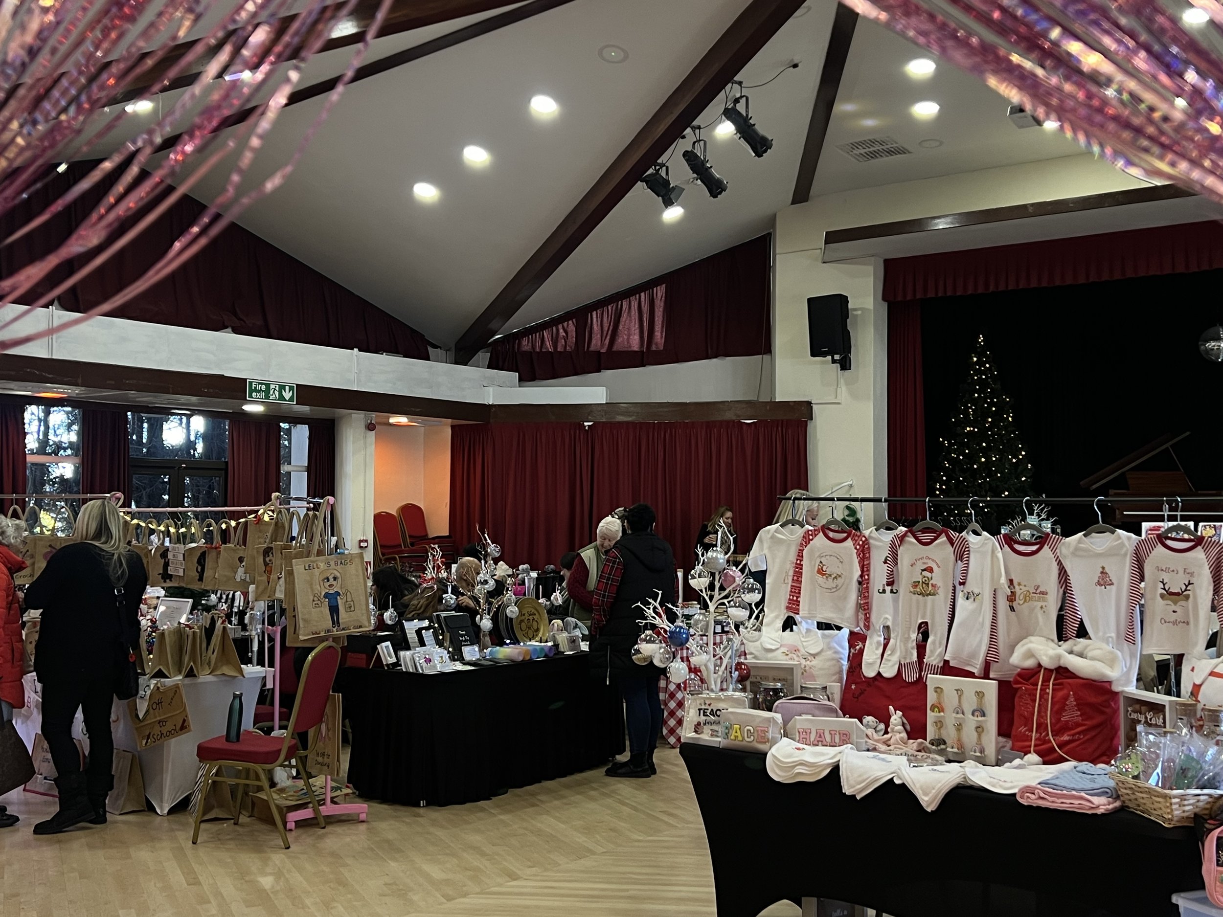 Indoor Christmas market or craft fair with holiday-themed crafts and decor on display, including Christmas sweaters, ornaments, and bags, in a room decorated for the holiday season.