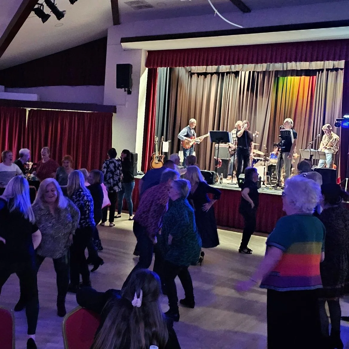 People dancing in front of a stage where a band is performing with guitars and microphones, in a dimly lit indoor venue.
