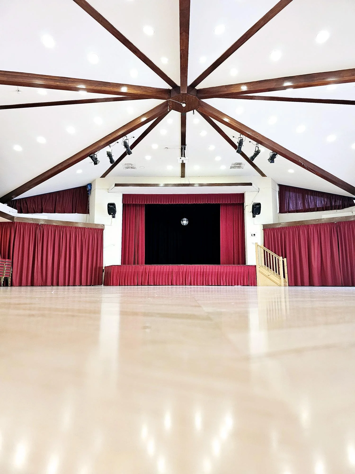 Empty community hall or event space with a stage, red curtains, and a wooden ceiling with beams.