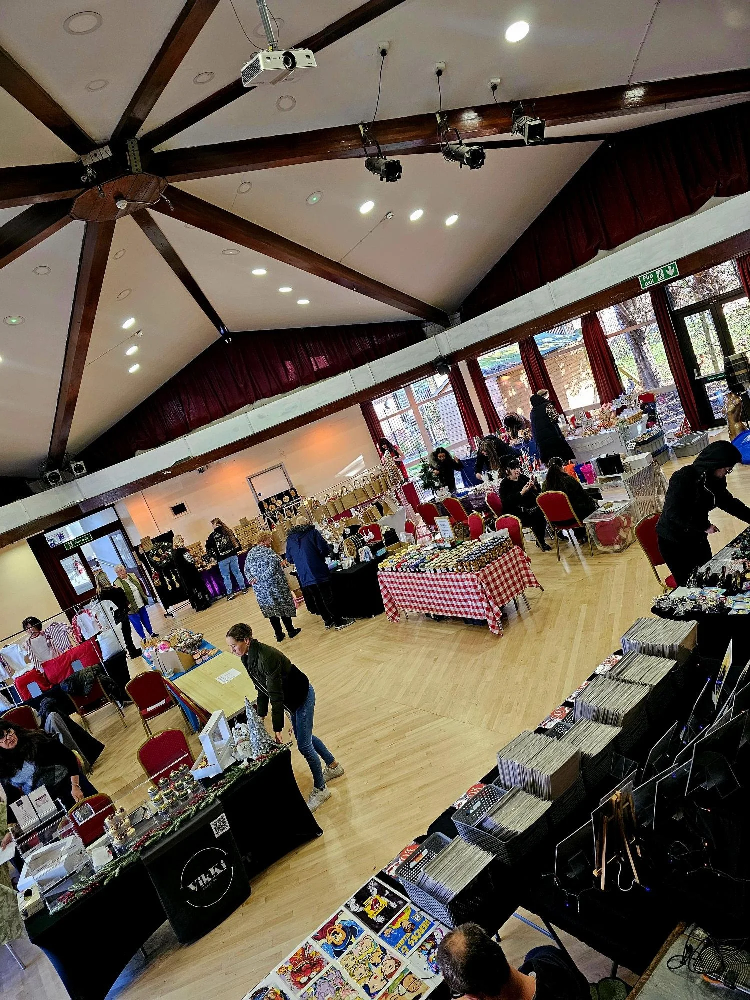 An indoor event space with booths and tables displaying various items for sale, including jewelry, crafts, and comics, with people browsing and walking around.