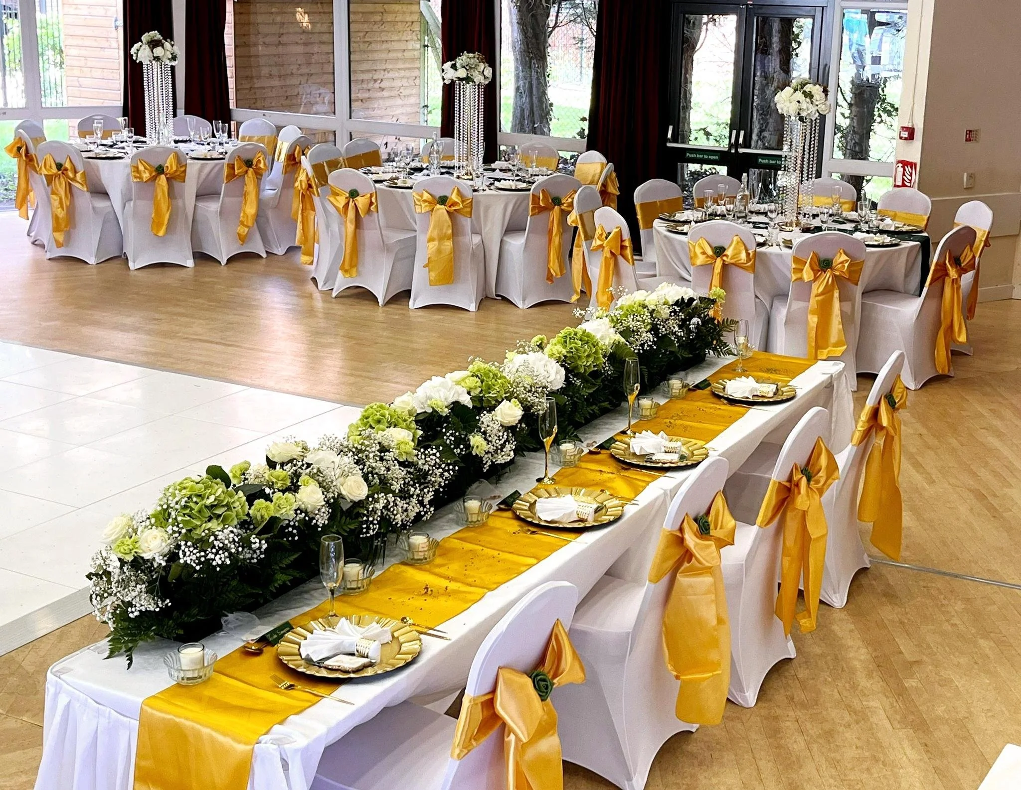 A banquet hall decorated for a special event with long tables covered in white cloths, gold table runners, and white chairs adorned with gold bows. The tables have floral centerpieces, glassware, and place settings, with a large floral arrangement ru