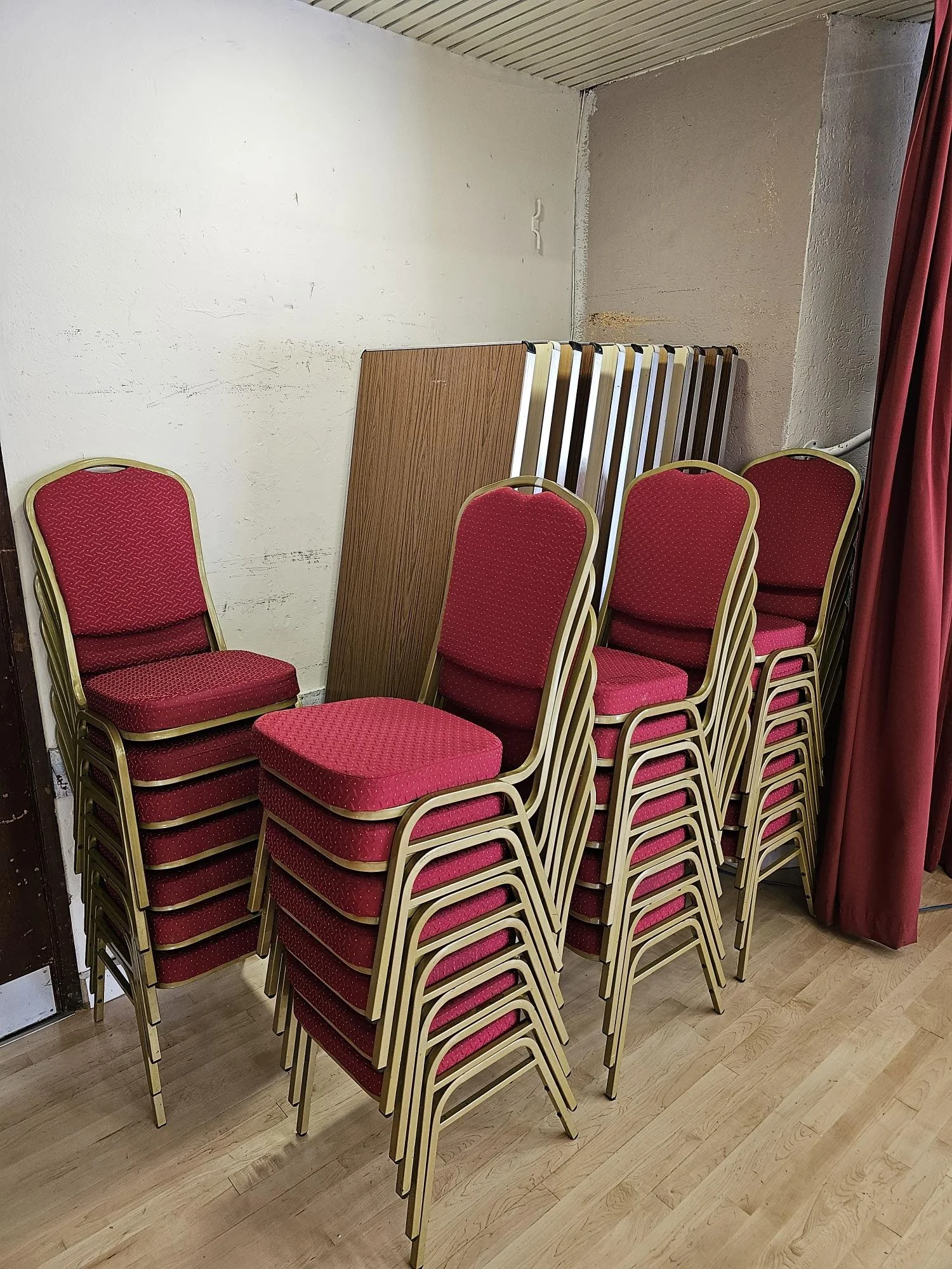 A stack of red upholstered chairs with gold frames stacked in a room corner, with some foldable tables leaning against the wall.