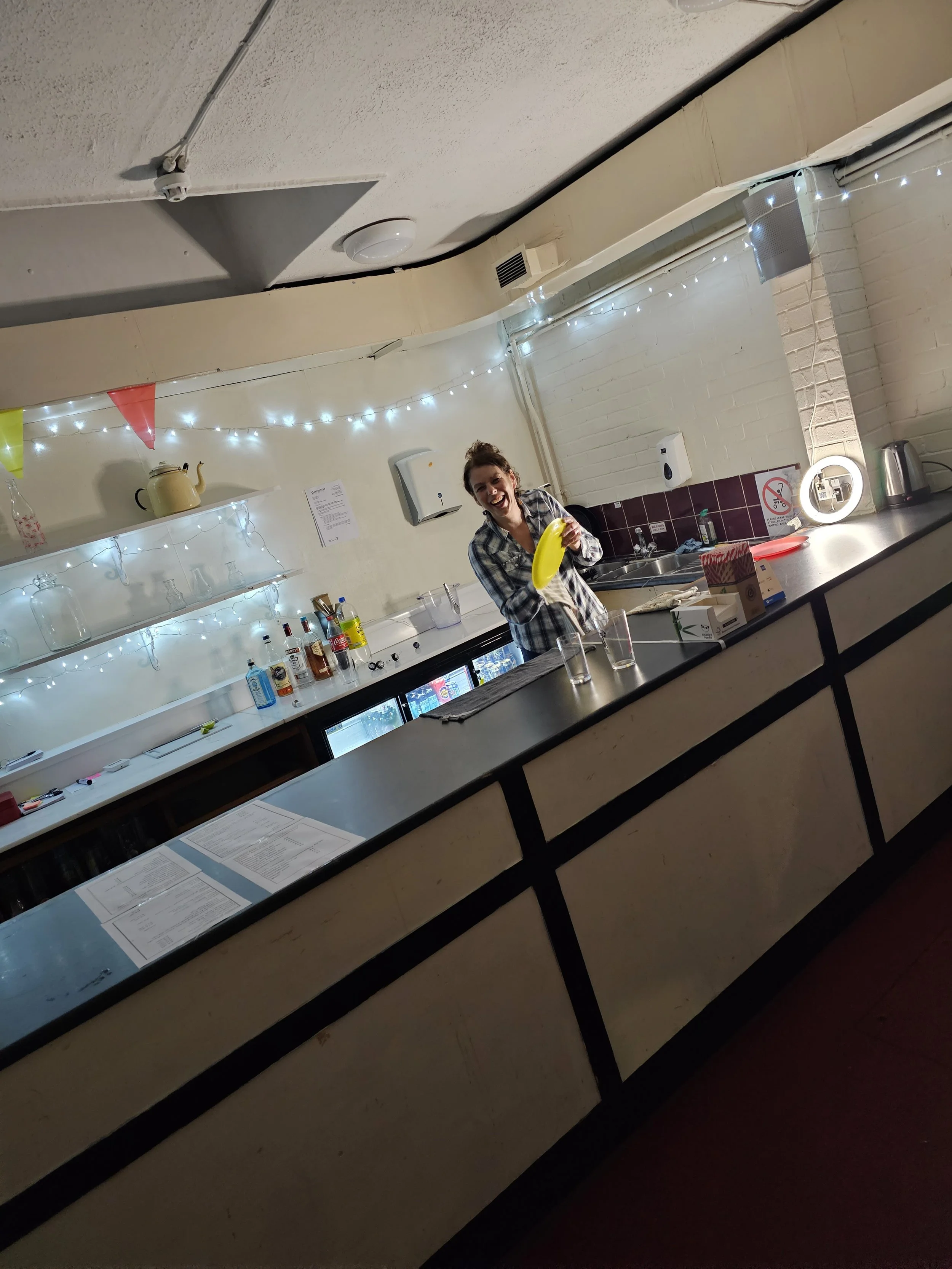 A woman smiling and holding a banana behind a bar counter decorated with string lights and colorful pennant banners, with bottles and glasses on the counter and shelves.