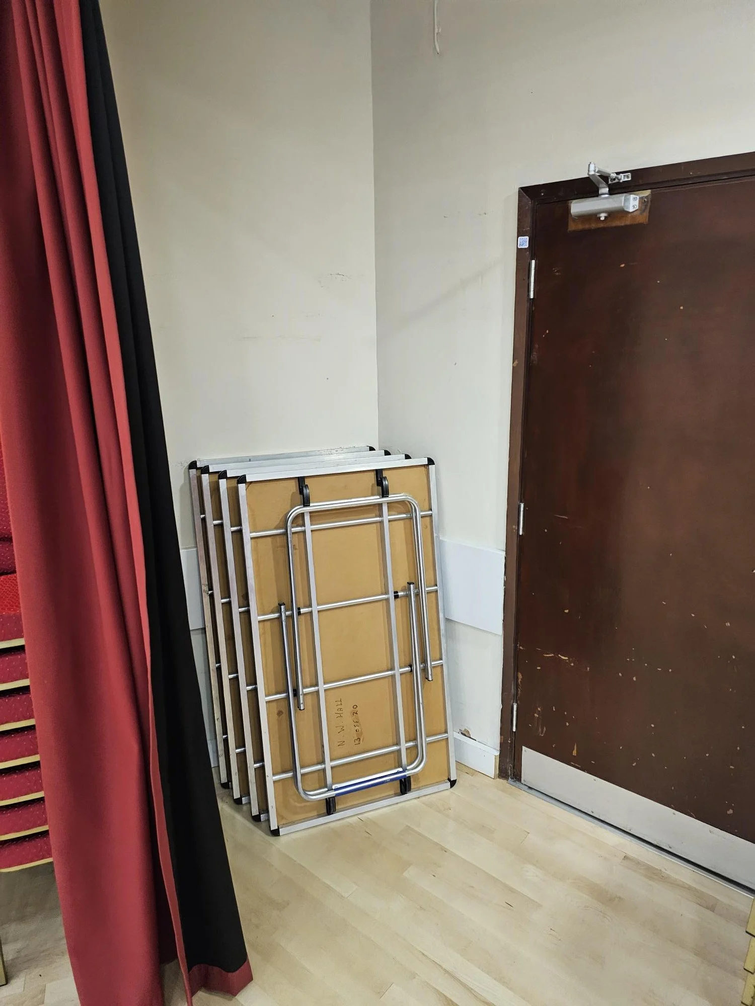 Folded tables and chairs stacked against a wall near a door in a room with a red curtain partially visible.