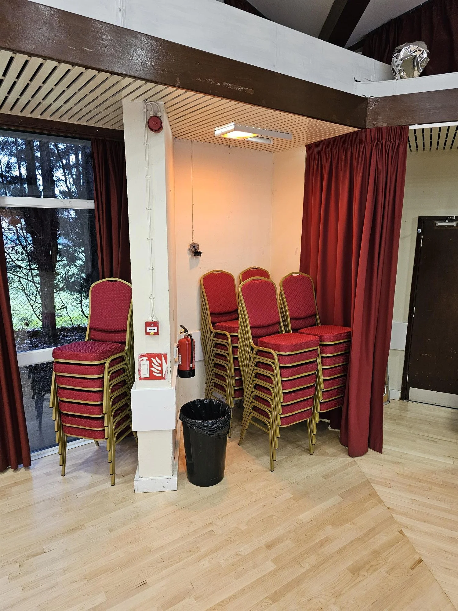 Stacked red chairs with gold frames next to a white pillar, near large window with outdoor trees, red curtains, fire alarm, fire extinguisher, trash can, and closed door in a room with light wood flooring.