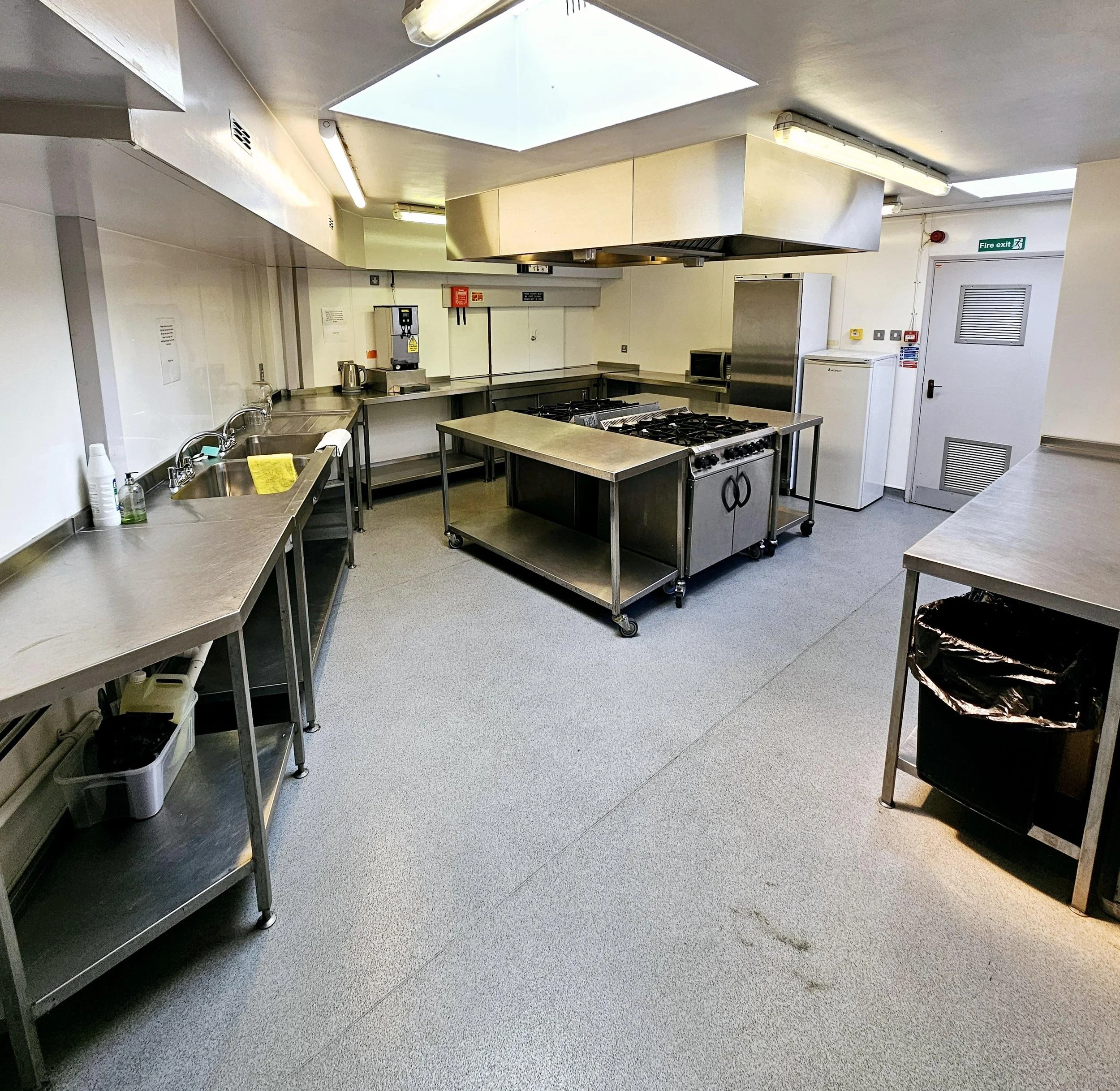 A commercial kitchen with stainless steel counters, a four-burner stove, a large refrigerator, a small microwave, a water boiler, and cleaning supplies. The room has a grey carpeted floor and white walls, with a window or skylight in the ceiling.