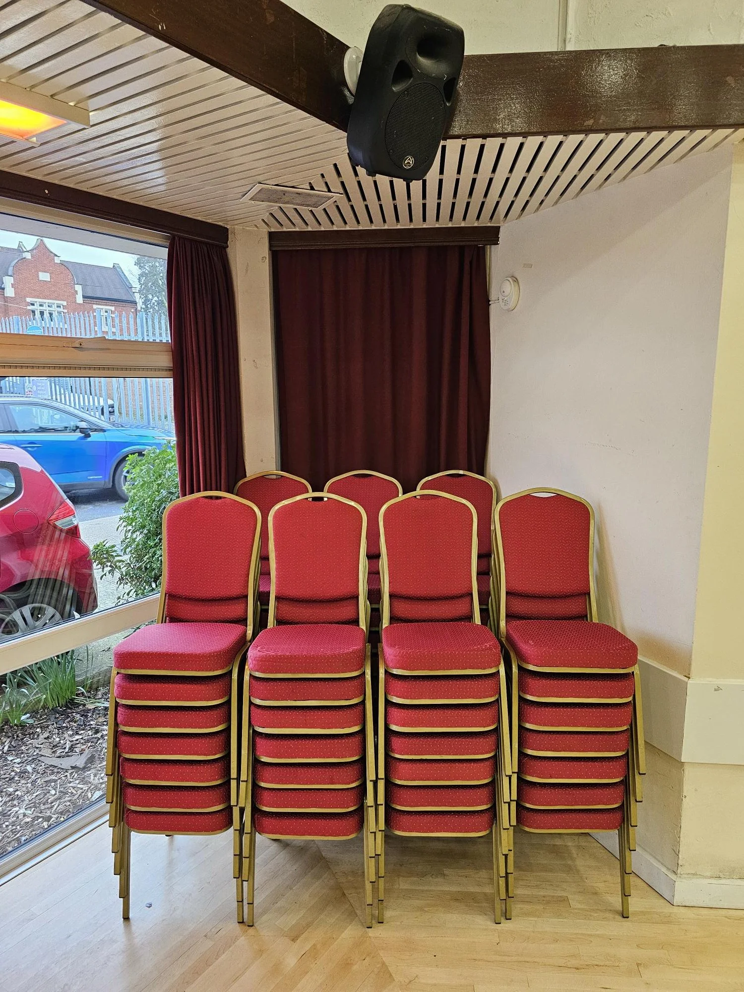 Stacked red cushioned chairs with gold frames arranged in front of a white wall and near a window, with red curtains and a black speaker mounted on the ceiling.