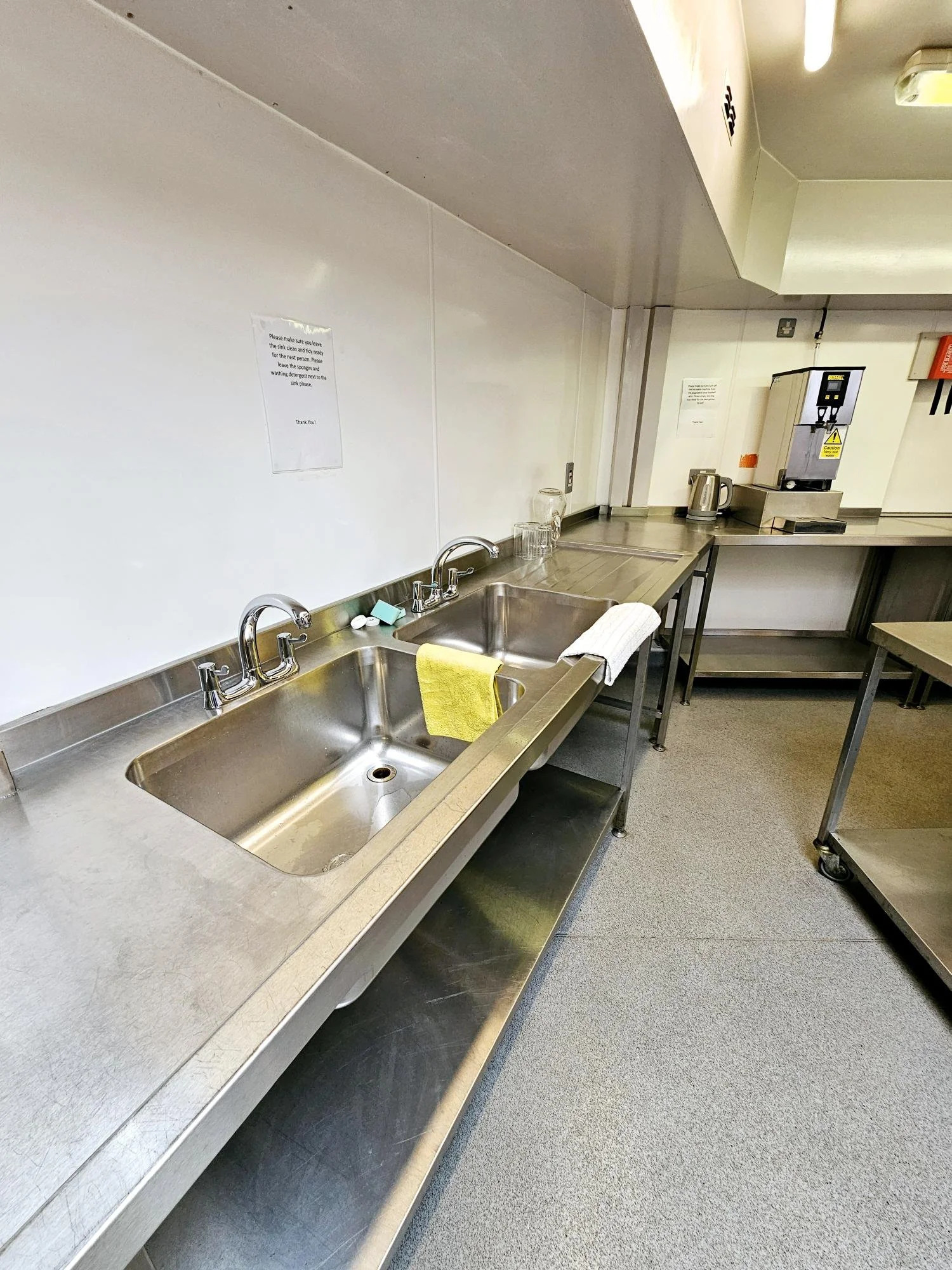 Stainless steel double kitchen sink with yellow and white towels, a soap dispenser, and glasses on the countertop. Behind, there are signs on the white wall, a silver electric kettle, and a commercial coffee machine. The room has beige carpeting.