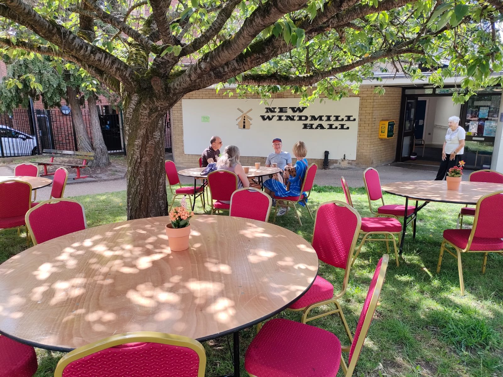 Outdoor patio with pink chairs and round tables, green grass, and a large tree providing shade. In the background, four people sit around one table, and a woman stands near the building entrance labeled 'New Windmill Hall.'