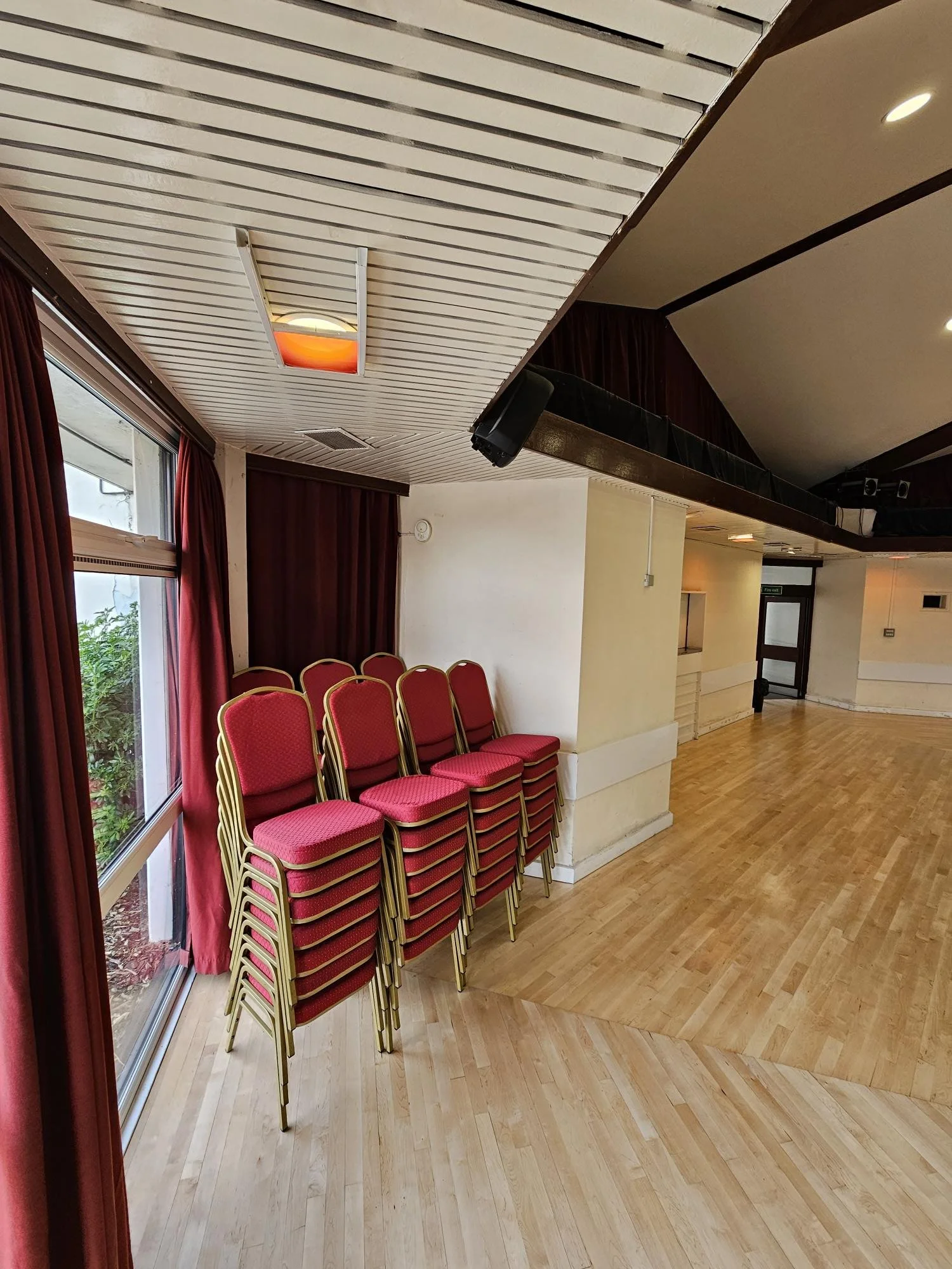 Stacked red banquet chairs with gold frames next to a window with red curtains in a room with wooden floors and white walls.