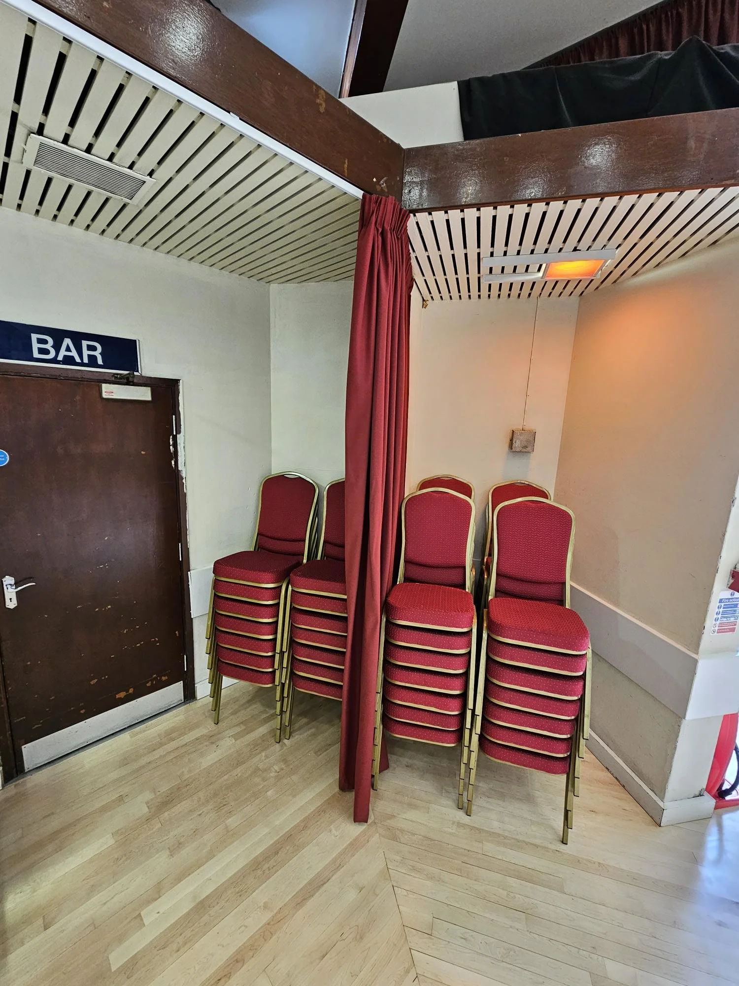 Stacked red chairs with gold frames against a white wall in a corner near a wooden door with a 'BAR' sign, separated by a red curtain, in a room with wood flooring and ceilings.