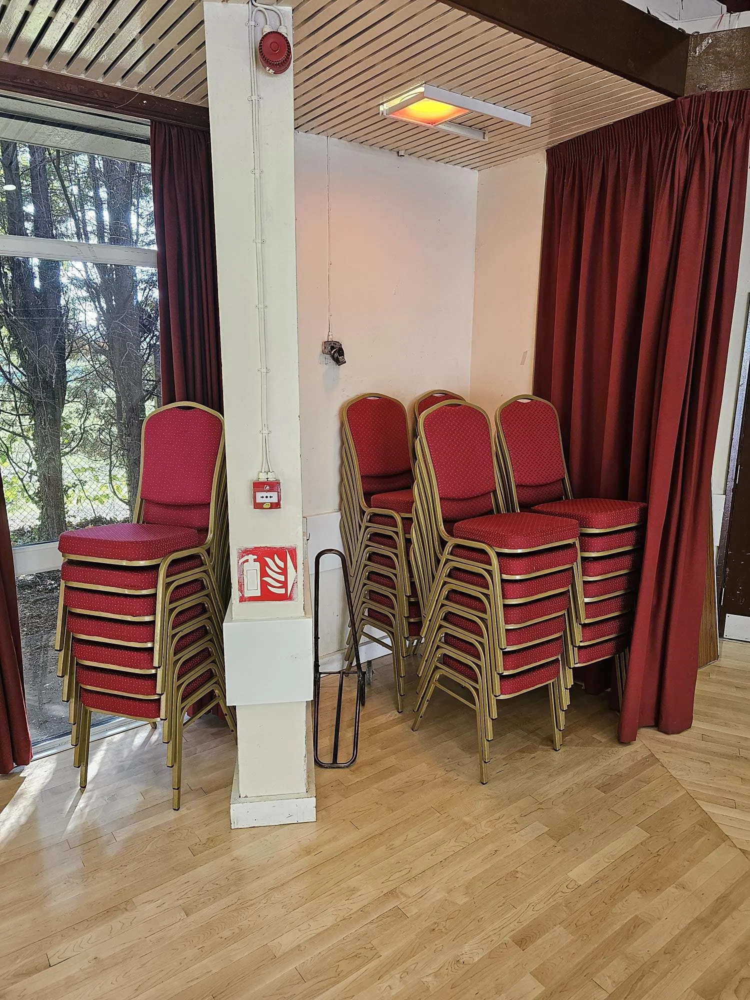 Stacked red chairs with gold frames in a room with red curtains and a window showing trees outside.