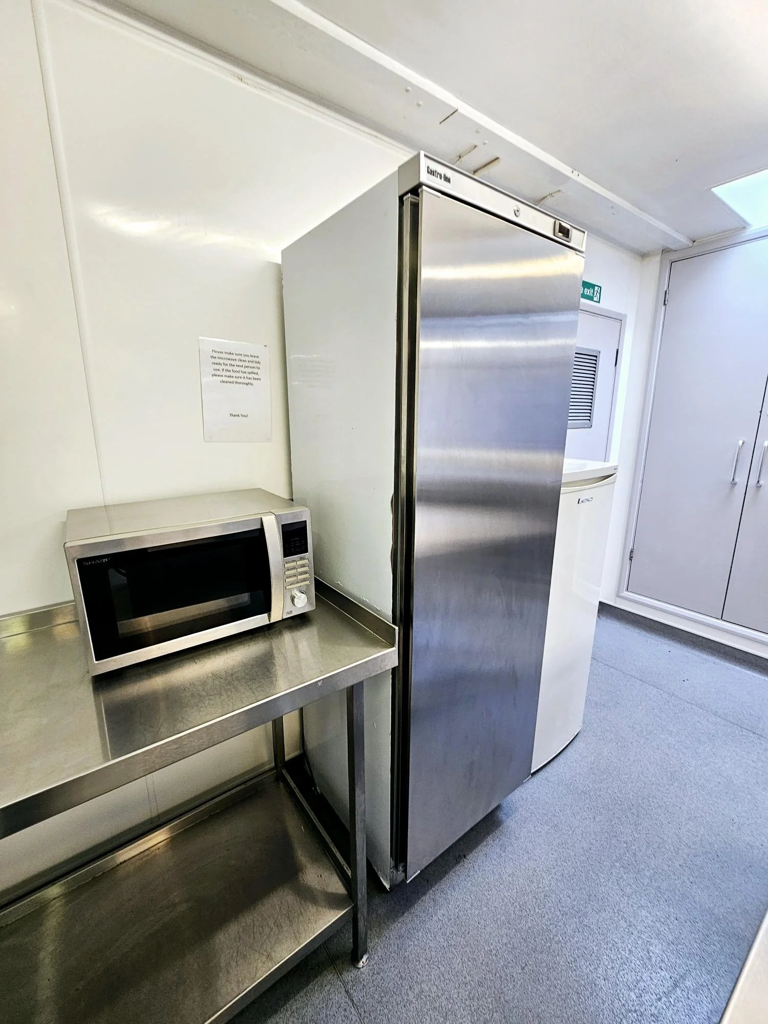 A microwave oven on a stainless steel cart next to a tall stainless steel commercial refrigerator in a kitchen or break room.