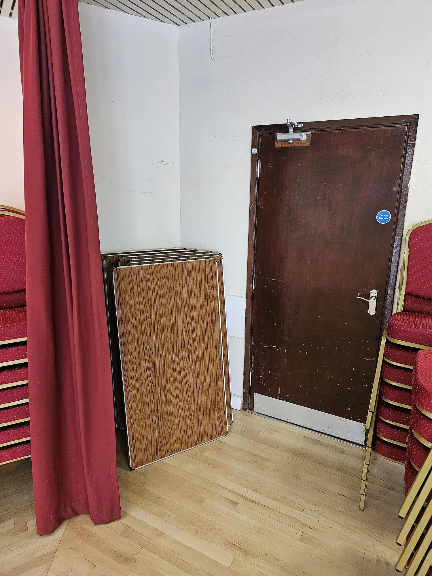 Stacked folding tables leaning against a corner wall near a closed wooden door, with red chairs stacked nearby and a pink curtain to the left.