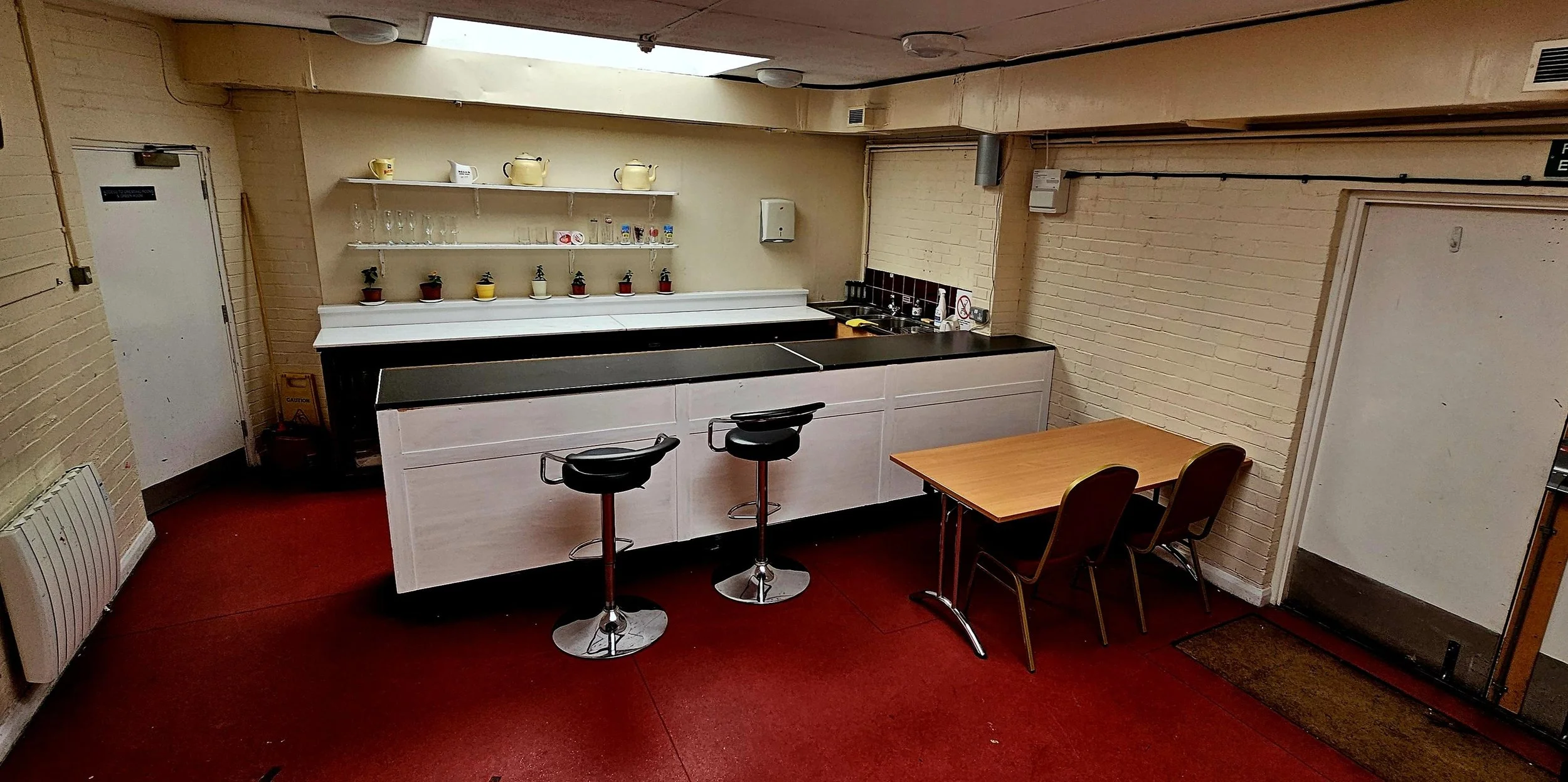 A small cafe or break room with a bar counter, two black bar stools, a small table with two chairs, and kitchen shelving with cups, teapots, and glassware. The room has beige brick walls, red carpeted floor, and a door on the right.