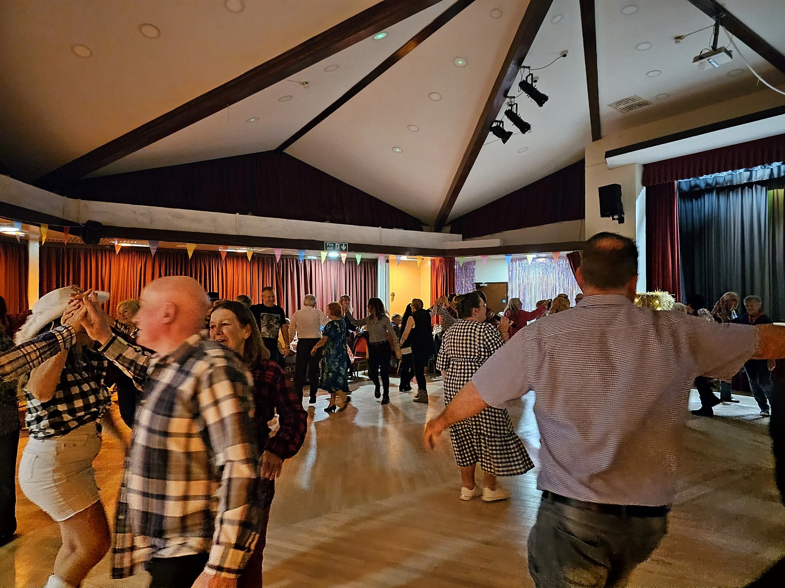 People dancing and socializing at a party or dance event in a decorated hall with colorful bunting and stage curtains.