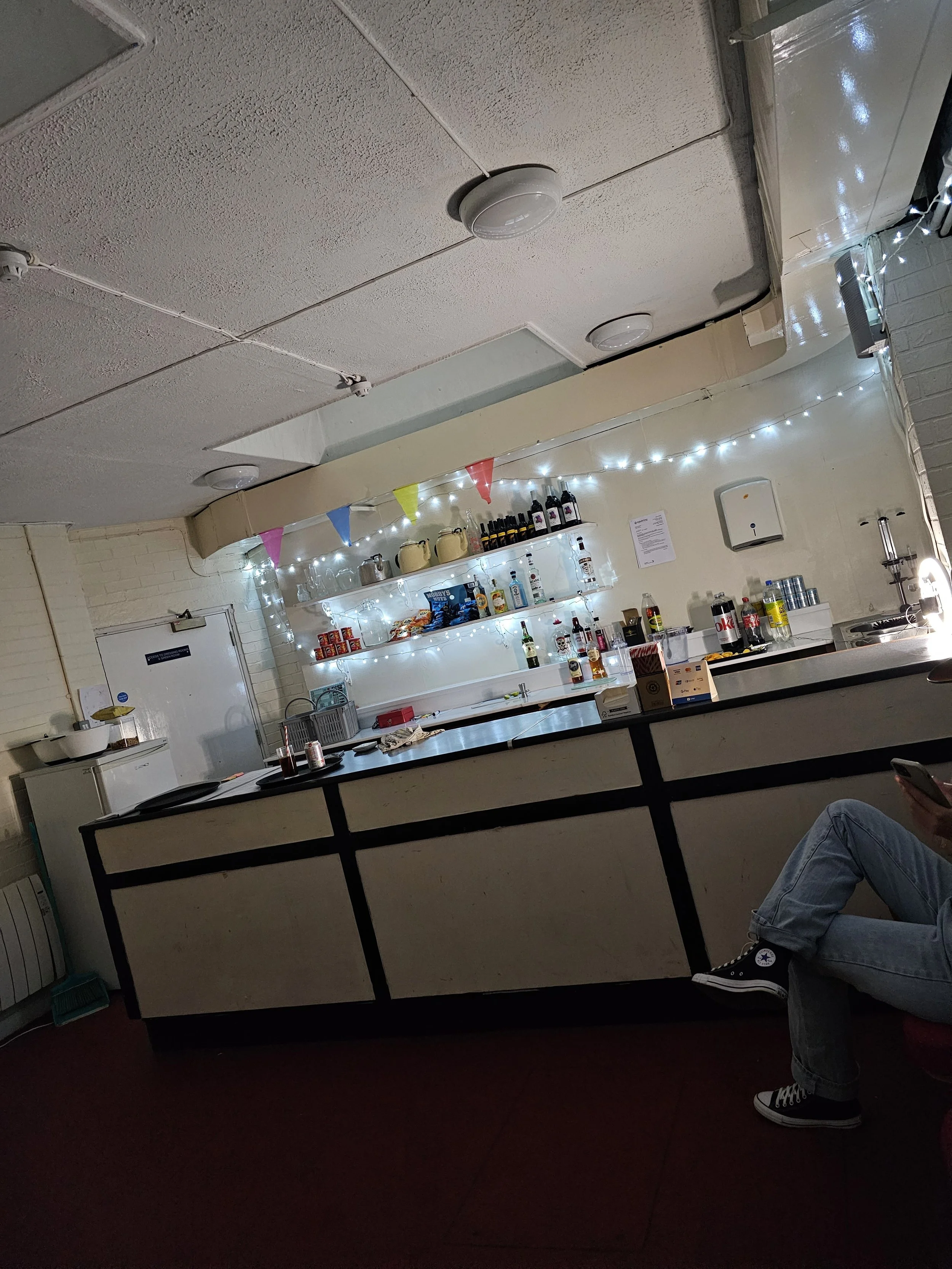 A bar area decorated with string lights and colorful flags. Shelves stocked with bottles, cans, and snacks are visible behind the counter. Part of a person sitting nearby, wearing jeans and Converse sneakers, is also in the image.