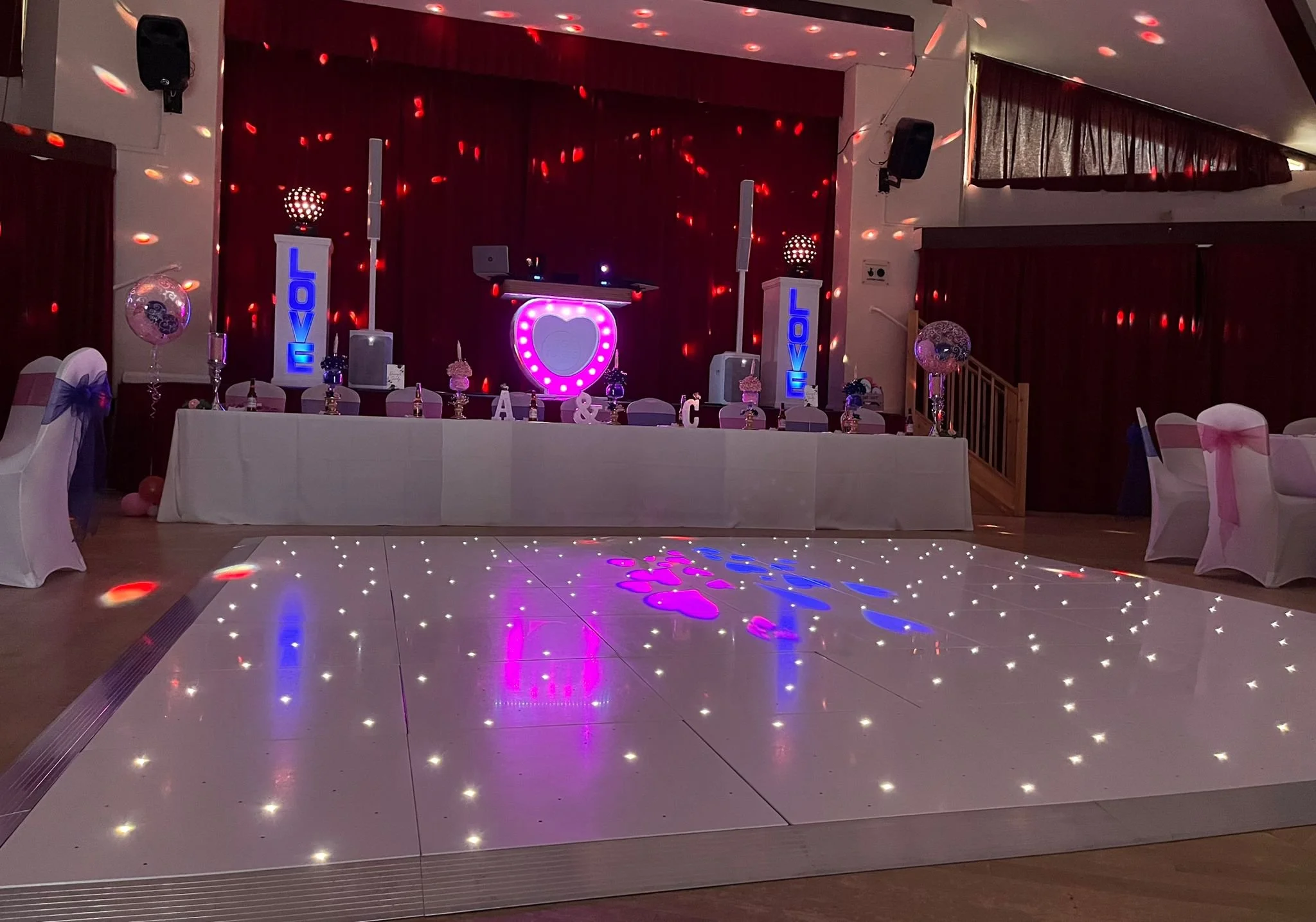 Wedding or party reception hall with decorated table and dance floor with lighting effects, featuring signs with the word 'LOVE' and a heart-shaped decoration behind the table.