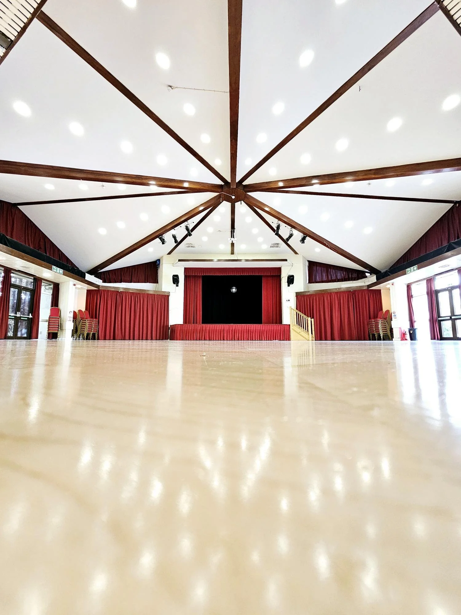 Empty event hall with polished wooden floor, red curtains, stage, and ceiling with spotlights.