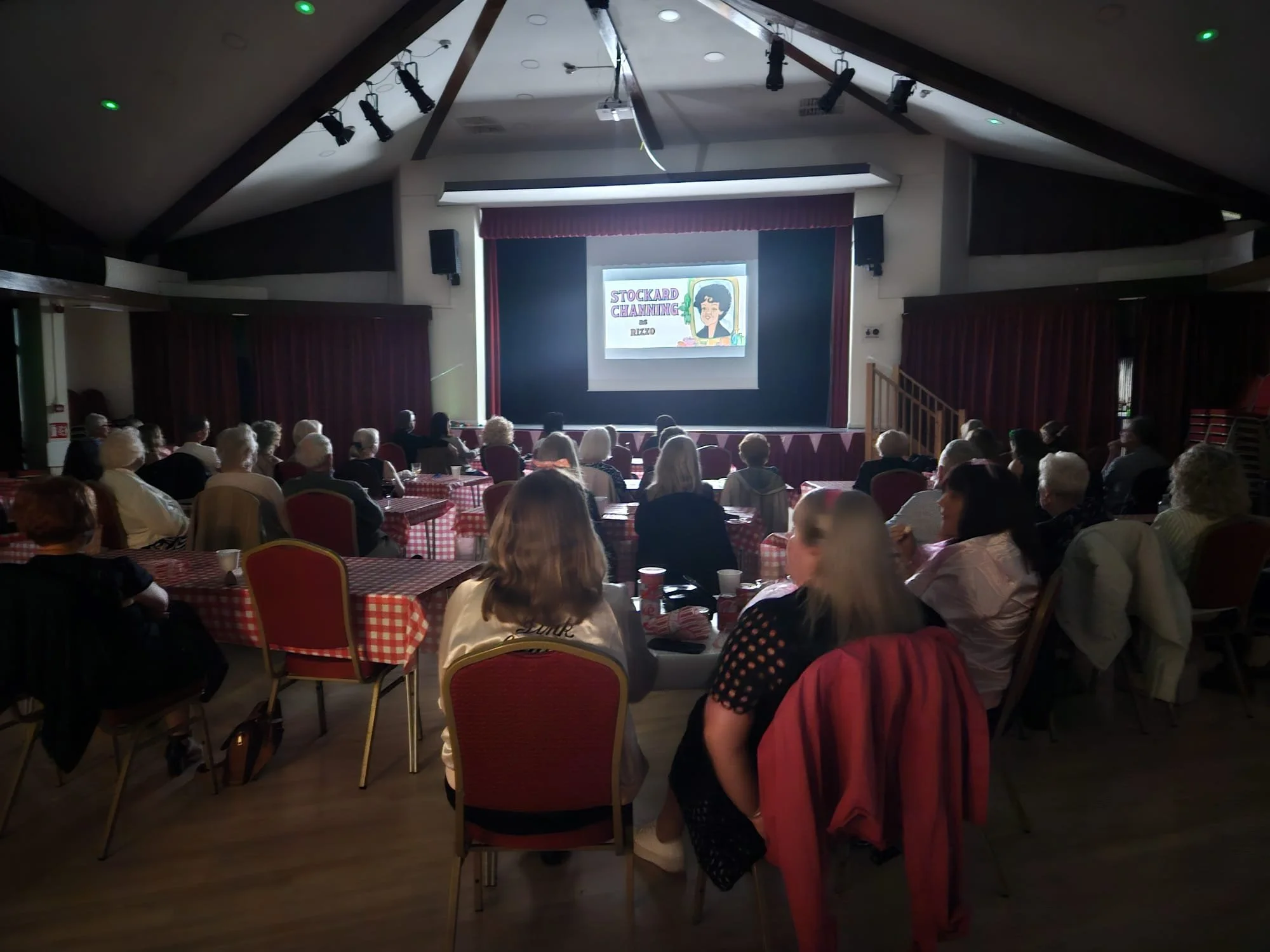 Audience watching a presentation in a large, dimly-lit auditorium with checkered tablecloths on tables and a stage with a projected screen.