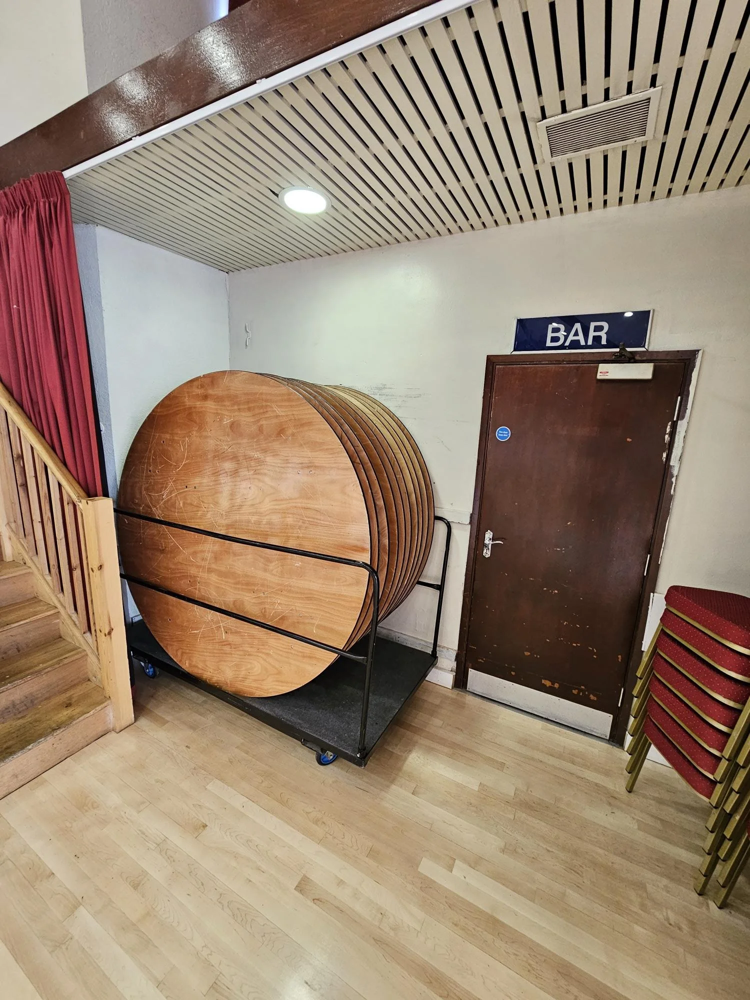 Stack of round wooden tables on a black rolling cart in a room near a brown door labeled 'BAR'.'}}#}