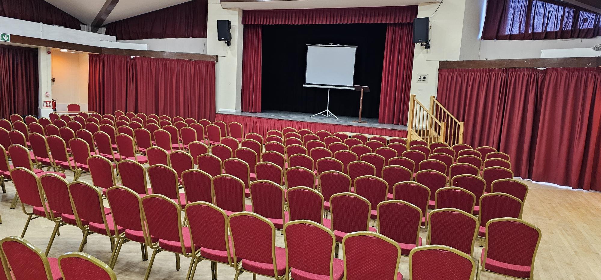 An empty auditorium with red chairs and a stage with a projector screen and red curtains.