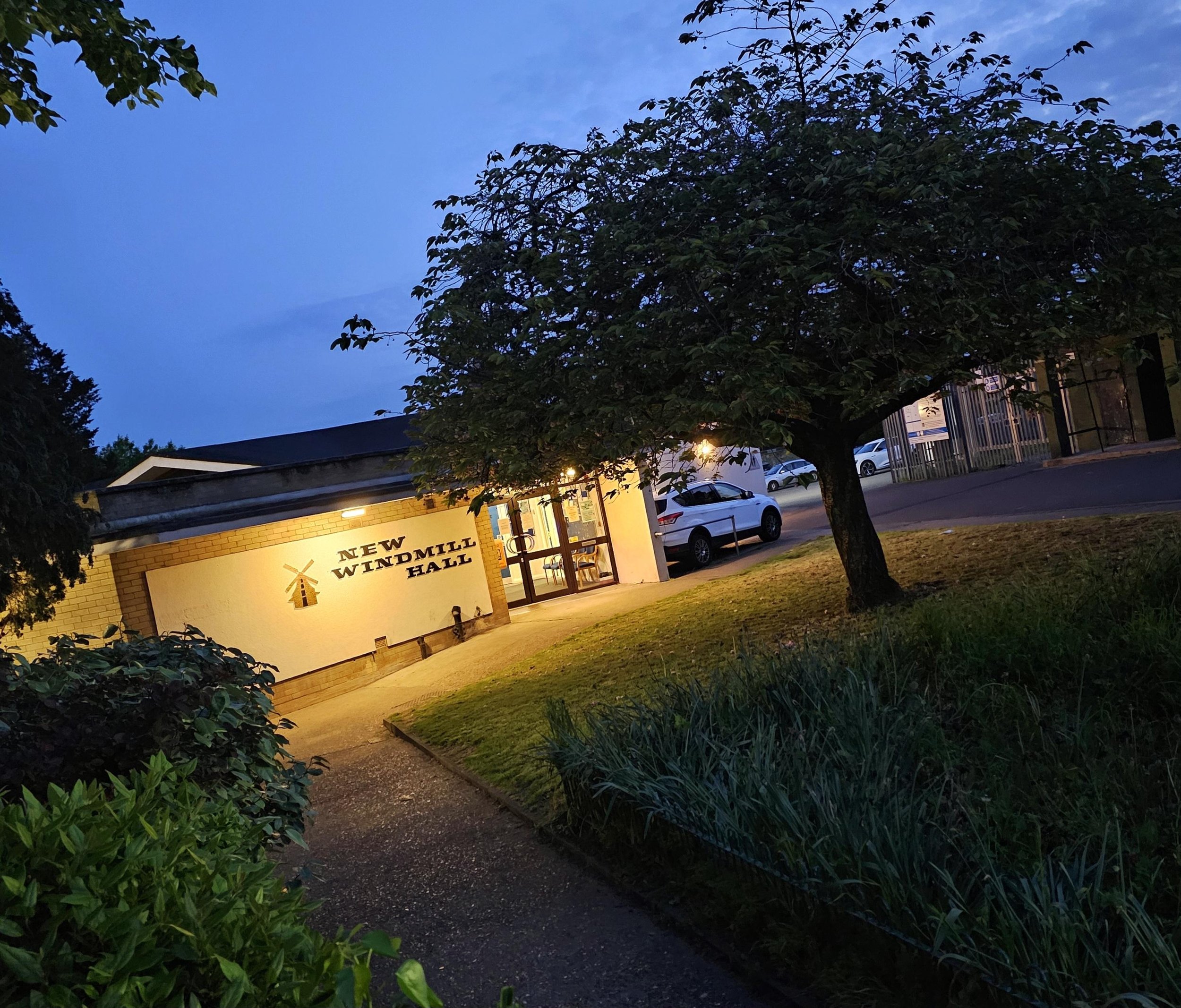 The entrance to New Windmill Hall, a building with a brick facade, illuminated by exterior lights, with a tree in front and cars parked nearby, during twilight.