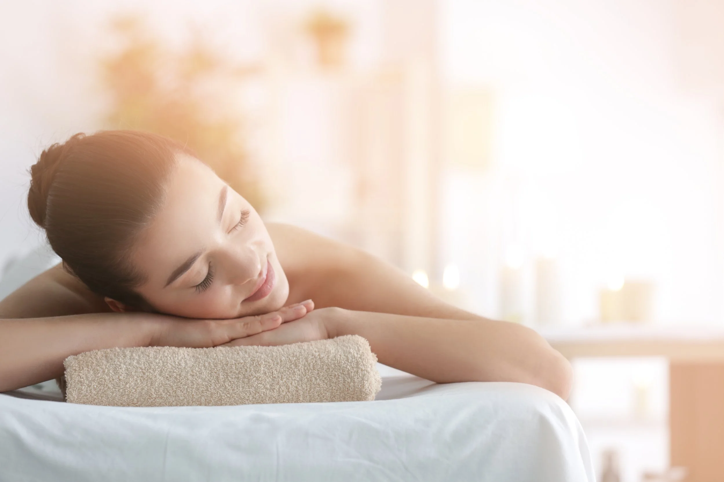 Young woman relaxing on massage table with eyes closed, receiving a massage or spa treatment, in a bright, softly lit room.