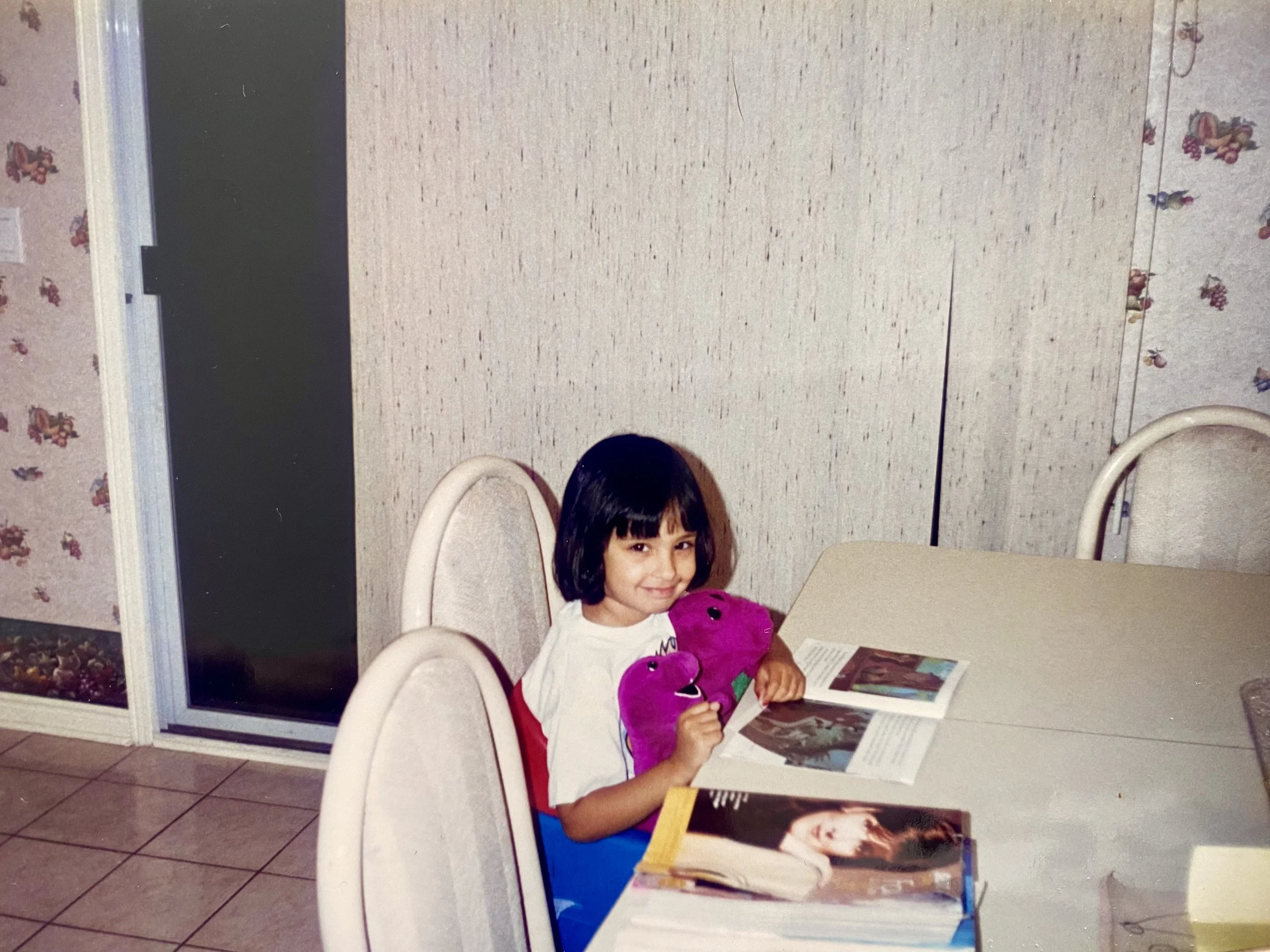 A young girl with dark hair and bangs sitting at a dining table, holding a pink plush toy, smiling at the camera, surrounded by open books.